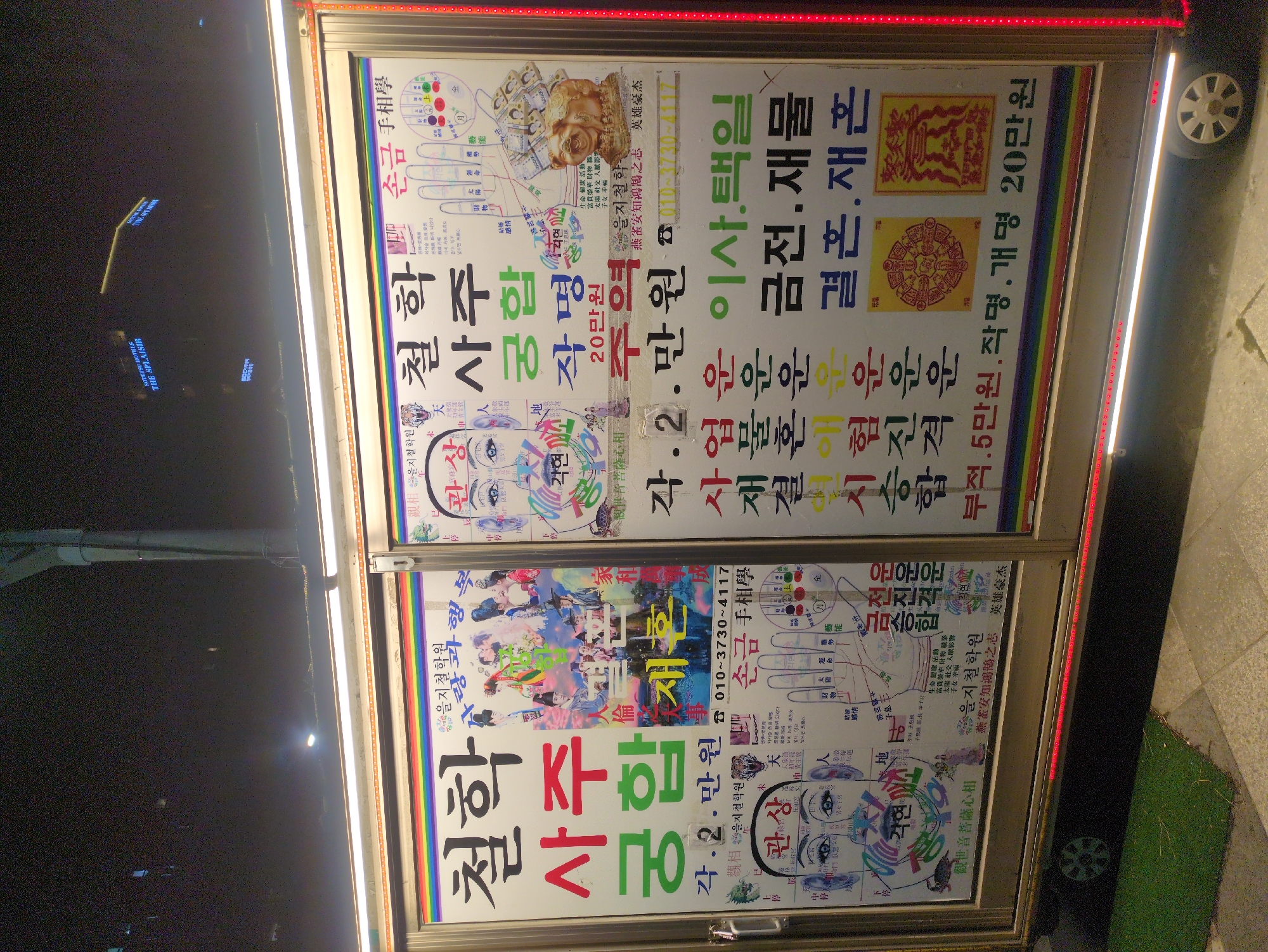 The image captures a mobile fortune-telling booth on a street, likely in Seoul, South Korea, judging by the Korean text. The booth features two large illuminated signs displaying information about palmistry, face reading, and compatibility readings. Details include prices (in Korean Won) for these services. The booth is equipped with wheels. The background shows night-time street lighting.
