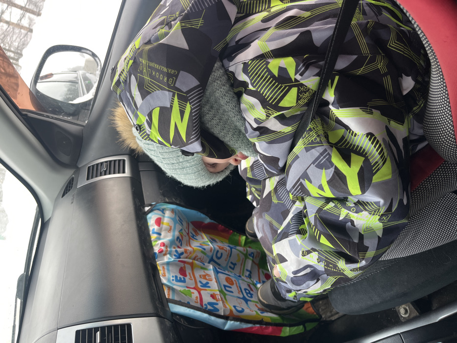 Within the front passenger seat of a vehicle, a young individual is observed in a curled, resting position. The exterior environment, visible through the windows and side mirror, is a snow-covered landscape under an overcast sky, with the location attributed to Италмас, Russia. The individual is dressed in a heavily insulated winter suit, predominantly black, grey, white, and neon yellow-green, featuring geometric patterns and the word "BROOKLYN". A light blue knitted beanie with a fur pom-pom covers the head, complemented by a matching knitted neck gaiter. Black boots are visible on their feet. The individual's feet are braced against the lower section of the dark grey dashboard, partially obscuring a colorful fabric organizer adorned with Cyrillic alphabet characters and illustrative images. A black seatbelt strap crosses the individual's chest. The vehicle interior shows dashboard air vents.