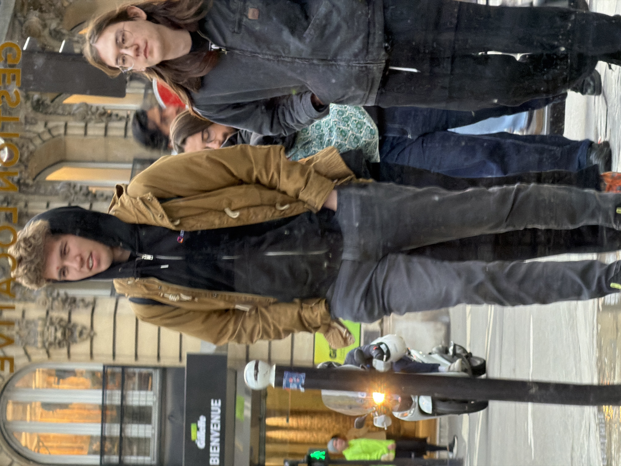 This image captures a street scene on a commercial street in Paris, France, under what appears to be overcast or diffused daylight conditions. The ground is wet, indicating recent precipitation.
In the immediate foreground, two young males are walking or standing. The male on the left has light brown, curly hair and wears a black hooded sweatshirt under a brown or tan jacket, with dark grey trousers. His hands are placed in his jacket pockets. The male on the right has long, dark brown hair, wears glasses, a dark grey or black zippered jacket over a dark shirt, and dark trousers with a visible white drawstring. His hands are also in his pockets. Both individuals are looking forward or slightly to their respective sides.
Behind the foreground subjects, several other individuals are visible. One person with dark hair and glasses is partially obscured, wearing a teal-colored patterned garment under a darker jacket and blue jeans. Further left in the background, a person in a bright neon yellow-green jacket and dark trousers stands near a pedestrian signal displaying a green 'walk' symbol.
A light-colored scooter with an illuminated headlight is present on the wet street in the mid-ground, with a rider wearing a white helmet. A tall black pole with a white spherical top and some stickers is prominent on the left side of the frame.
The background features a multi-story building with classical Parisian architecture, characterized by light-colored stone, arched windows with dark railings, and decorative carvings. Reflections in a large window in the upper background partially show the text "L'OCCITANE" and "GESTION" (likely "Gestion Locative" or similar). A sign on the building's lower left displays "Gillette BIENVENUE".
