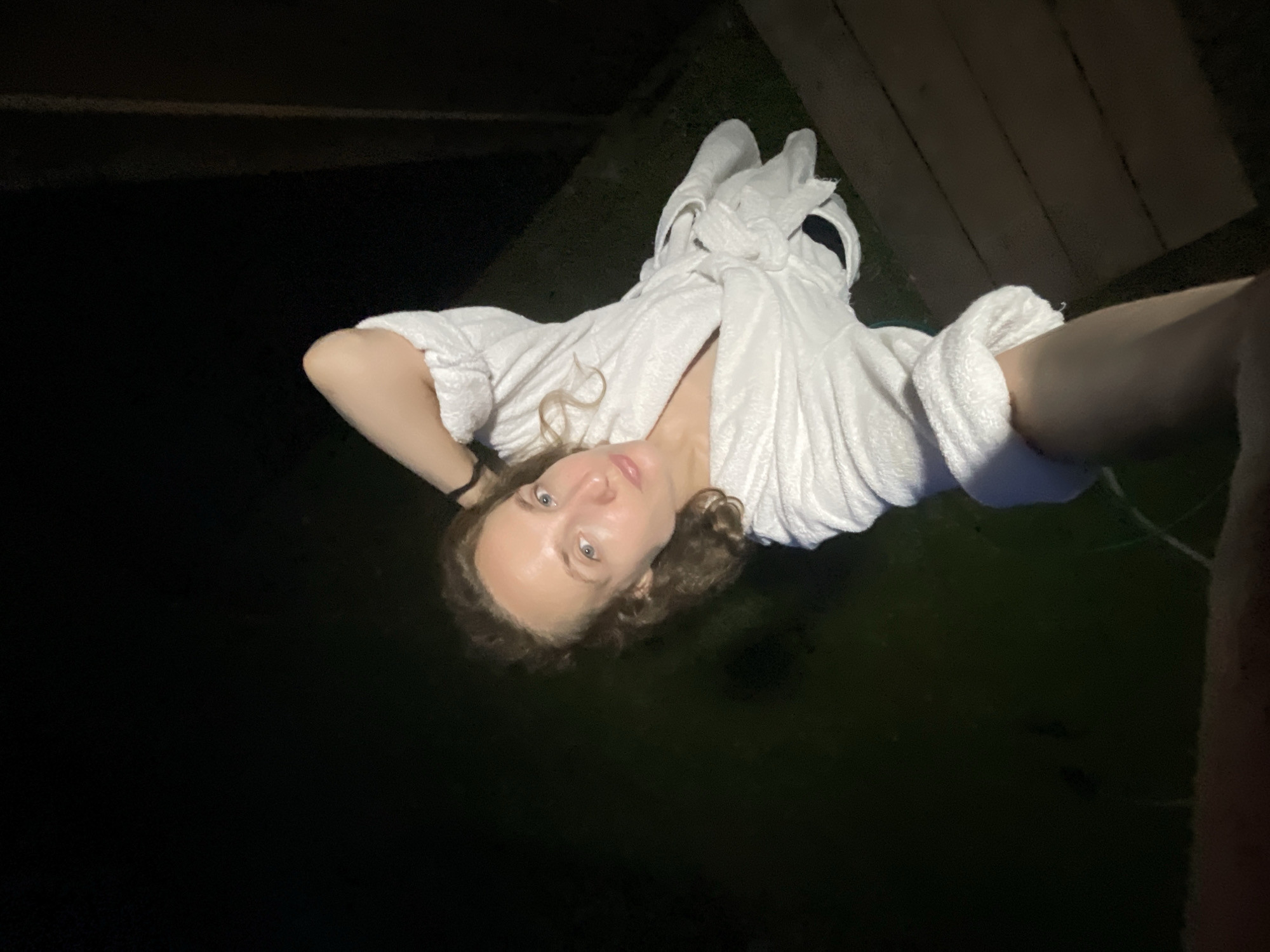 A woman, wearing a white bathrobe, is lying in dark water. Her head is tilted back, and her arms are bent at the elbows, with her hands resting behind her head. She has light brown curly hair, blue eyes, and light skin. The bathrobe is cinched at the waist and the sleeves are rolled up. The background is dark, suggesting it is nighttime. To the right of the woman, there are wooden planks, possibly part of a dock or deck. The water appears calm and dark, with some subtle ripples visible. The overall mood of the image is relaxed and serene, perhaps indicative of a quiet moment of self-care. There is no visible text in the image.