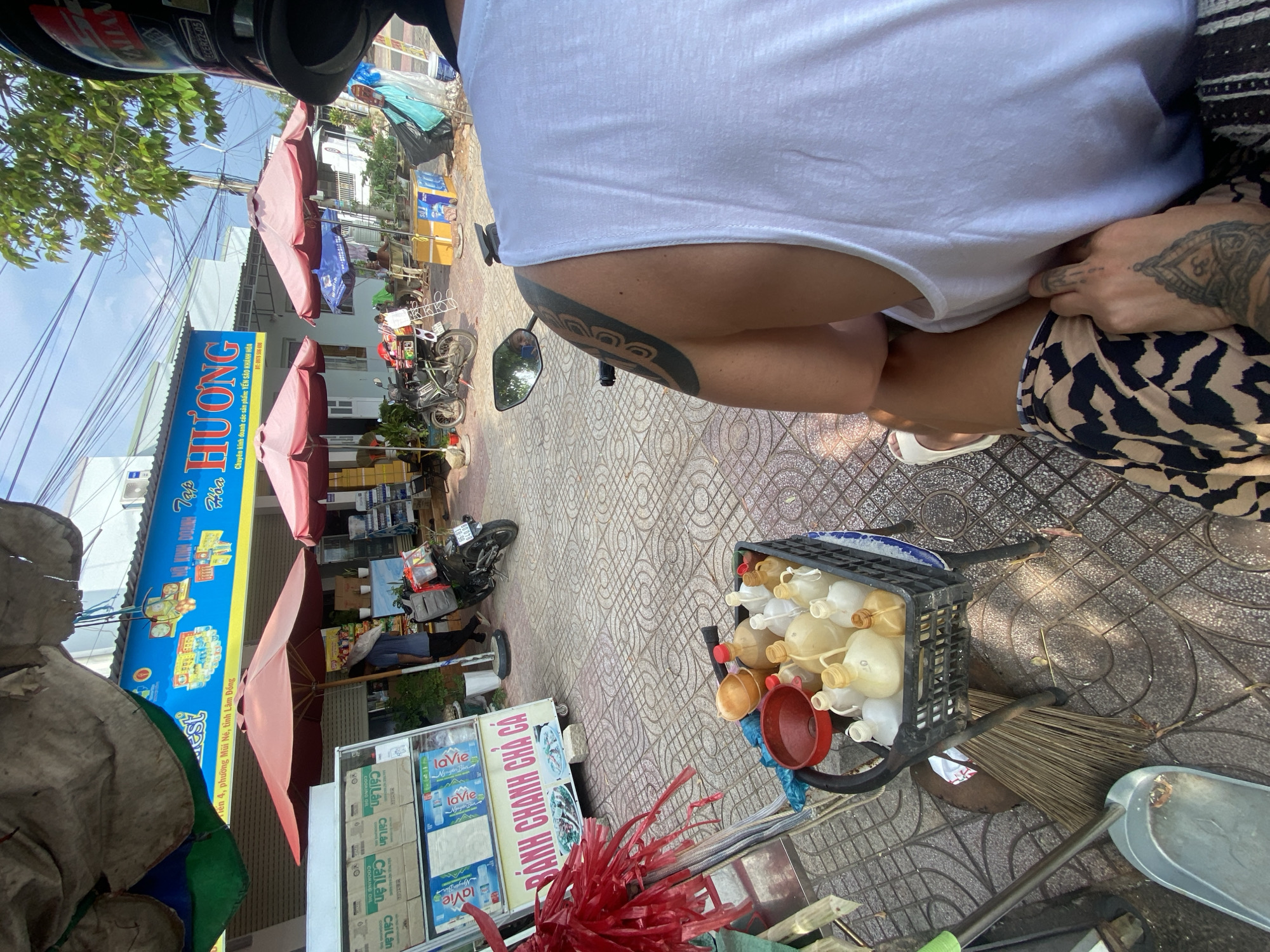 The image shows a street scene in Mũi Né, Vietnam. In the foreground, a person is seated on a motorcycle, viewed from behind. They are wearing a white tank top and patterned shorts, with tattoos visible on their arm and hand. To the left, a crate filled with various plastic bottles and a red funnel sits on a stand, with a broom and dustpan nearby. Behind this, a refrigerator display case is stocked with bottled drinks. In the background, a storefront with a blue awning displaying the word "HUONG" is visible, along with several red umbrellas shading outdoor seating areas. Motorcycles are parked nearby, and a person is walking past the storefront. The street is paved with patterned tiles.