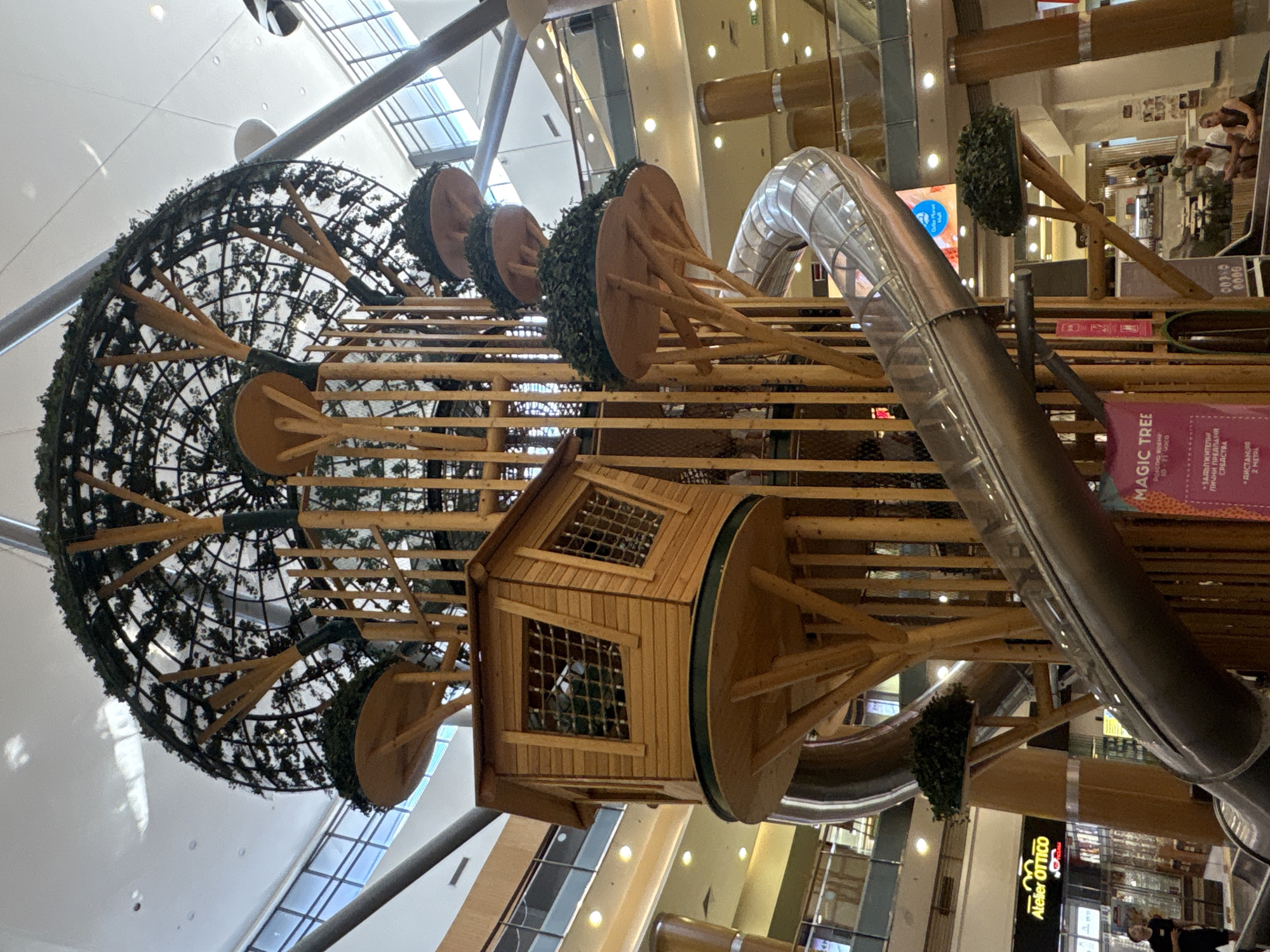 The image shows a large, indoor play structure designed to resemble a tree. The structure is made of wood and features multiple levels with platforms, a wooden playhouse with windows, and a shiny metal slide that winds down from one of the upper levels. The top of the structure is covered with a dark green, mesh-like material adorned with artificial foliage, creating a canopy effect. Wooden poles support the various platforms, and artificial greenery is strategically placed around the structure. In the background, the interior of a shopping mall is visible, with multiple floors, escalators, shops, and lighting fixtures. A sign at the base of the structure reads "MAGIC TREE". A few people are visible in the background, seated and standing. The scene is well-lit, suggesting it is daytime or the mall is brightly illuminated.