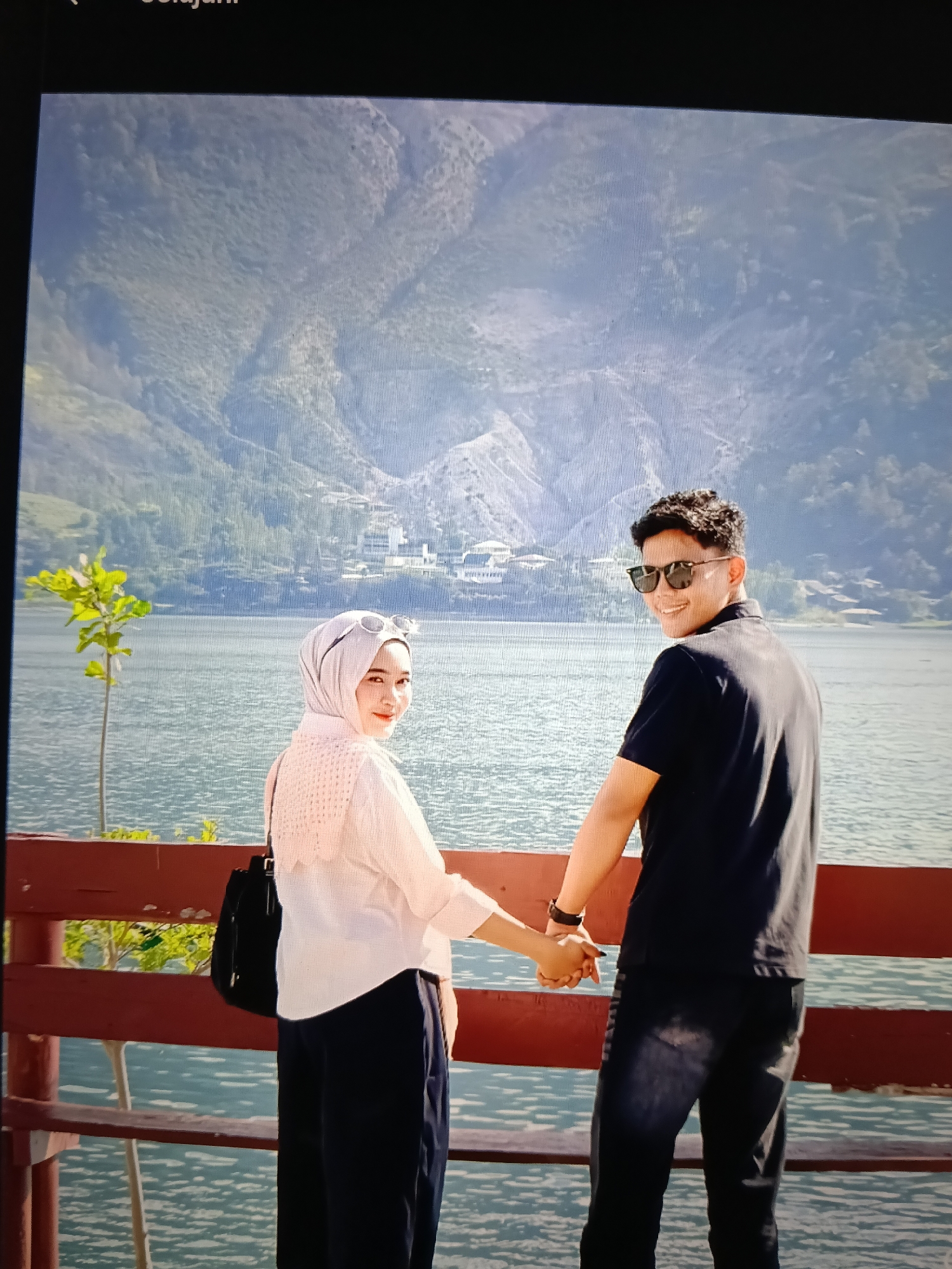 An outdoor, waist-up, medium shot shows a young adult couple holding hands and standing by a body of water. The man is on the right and the woman is on the left. They are both looking away from the camera. The woman is wearing a light-colored hijab and a light-colored blouse, and the man is wearing a dark-colored polo shirt and jeans. They are standing on a wooden railing with a lake or ocean and mountains in the background. The lighting is natural and the image has a slight greenish cast. The location is identified as the city of Raha, Indonesia, specifically the waterfront.