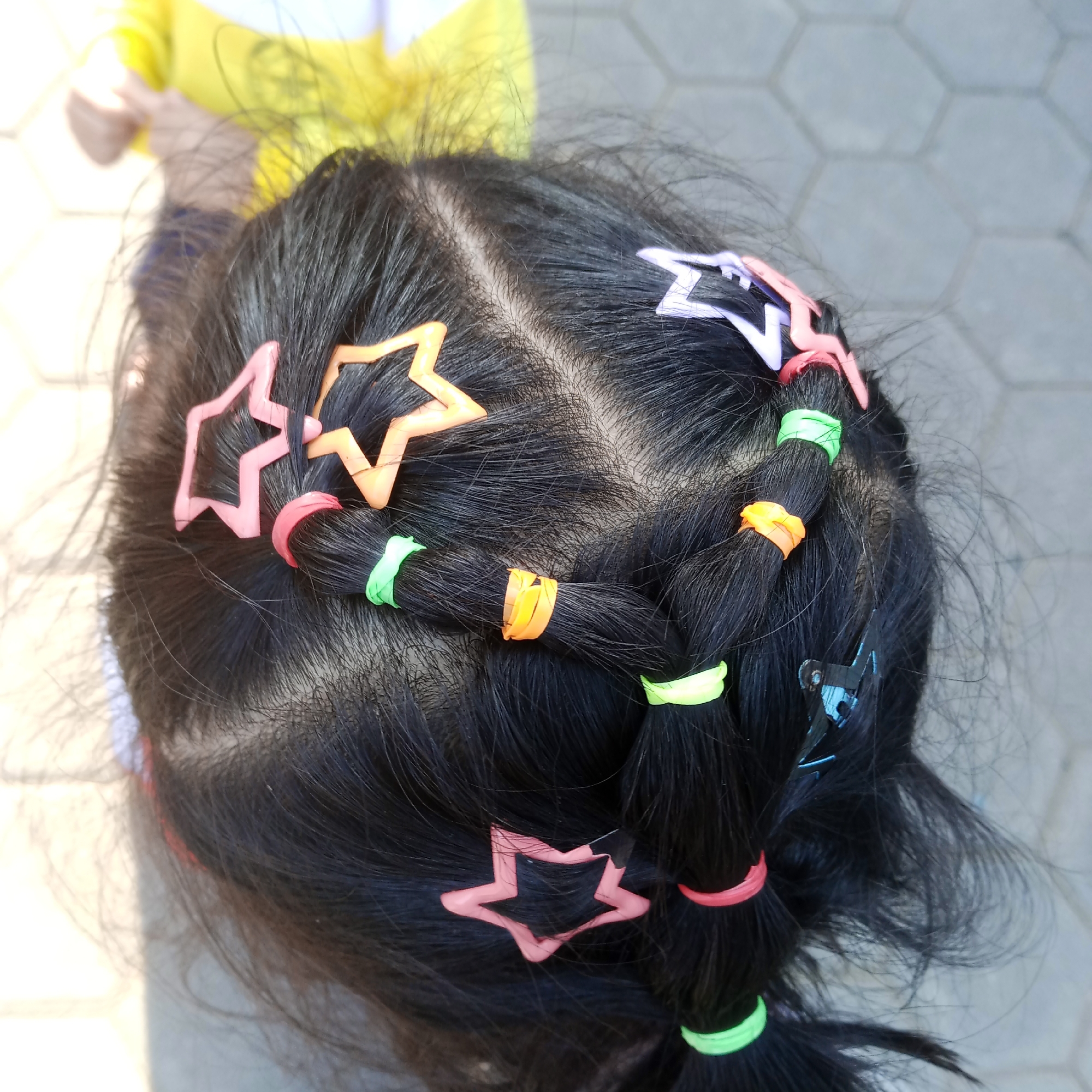 The image shows a close-up, overhead view of the back of a child's head, focusing on their black hair. The hair is styled into braids, secured with colorful hair ties in orange, green, and pink. Several star-shaped hair clips in pink and orange are also visible, decorating the hair. A blue hair clip with a metallic sheen is present in the lower right portion of the frame. The background consists of a light-colored, hexagonal tiled surface, suggesting an outdoor setting. The child is wearing a yellow garment, parts of which are visible in the upper left of the image. The lighting suggests it is daytime. The scene is devoid of any contextual markers that would indicate a specific city or location like Rejosari, Indonesia.