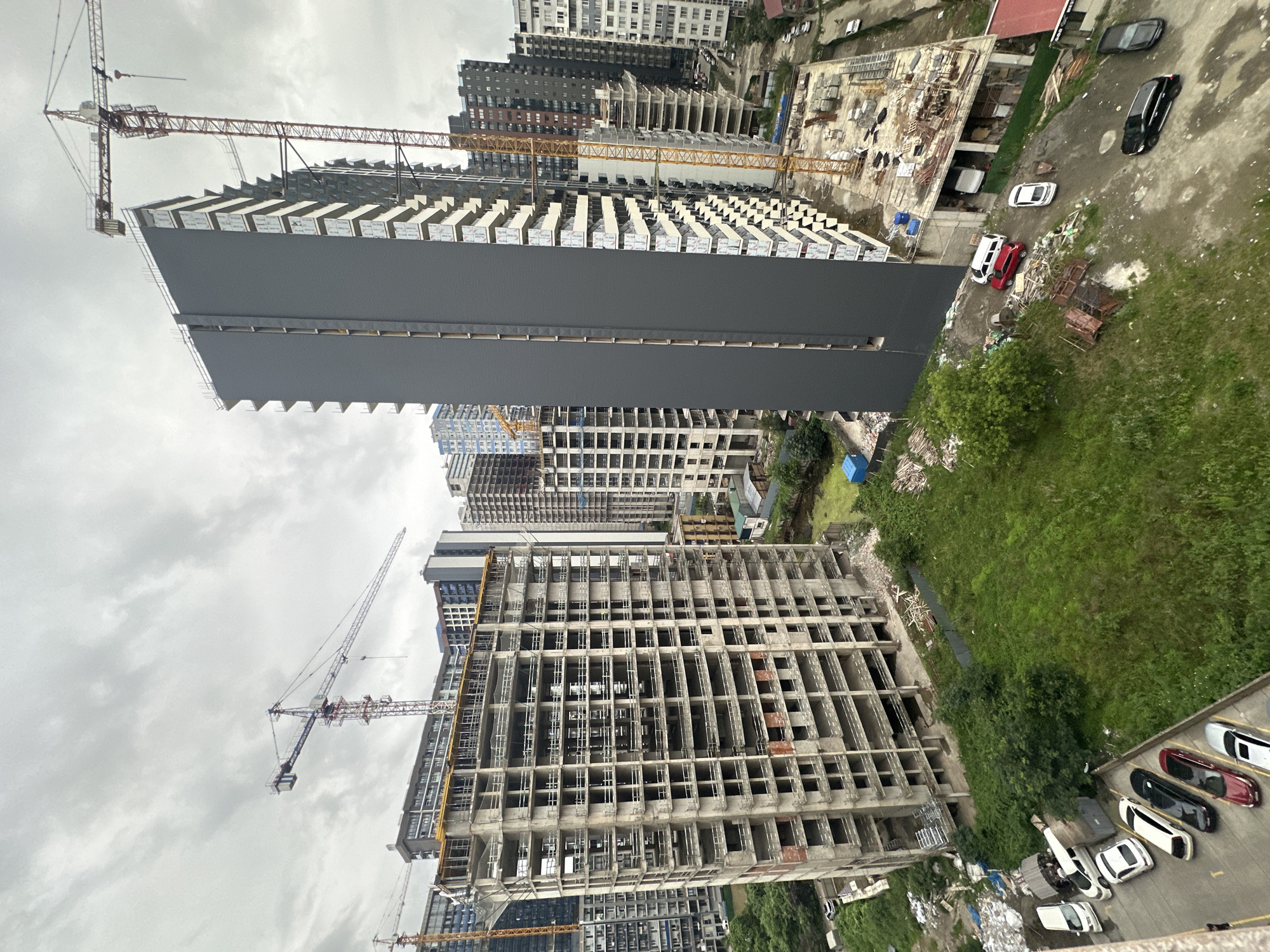 The image depicts a construction site in Batumi, Georgia, under a cloudy sky. Dominating the foreground is a partially constructed, multi-story building made of concrete, with visible rebar and open floors. To the right, a tall, dark gray, completed building stands next to a yellow construction crane. Further in the background, more high-rise buildings, some under construction and some completed, are visible, interspersed with green trees and patches of unkempt grass. Several vehicles, including white cars, a red car, and a black SUV, are parked in a lot to the left and on a nearby road. Construction materials like wood planks and debris are scattered around the site. The scene conveys an ongoing urban development project in a coastal city.
