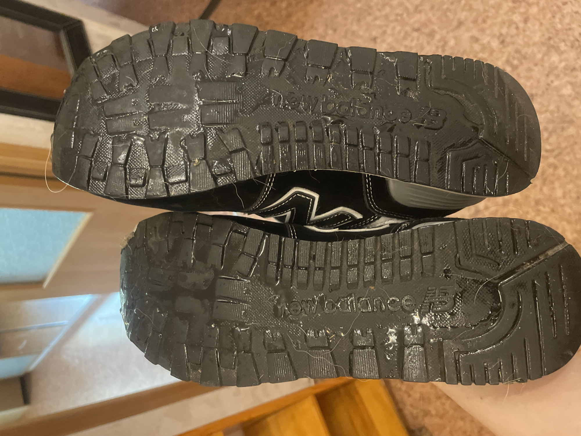 This image shows the soles of two black New Balance sneakers. The soles have a patterned tread and appear to be wet and dirty, with some white hairs stuck to them. One of the soles has the "New Balance" logo embossed on it. The sneakers are being held by a person, whose hand is visible in the lower left corner of the frame. The background suggests an indoor environment with wooden doors and what appears to be a hallway. There is no indication that this scene is located in Saint Petersburg, Russia.