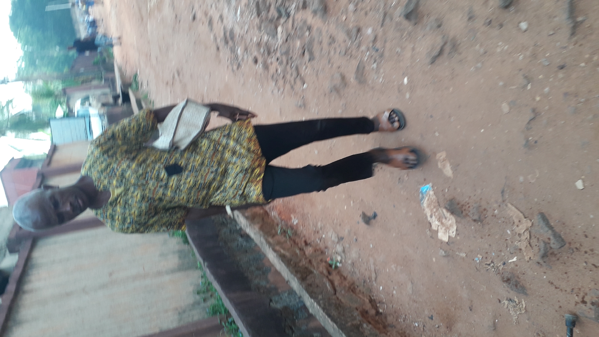 An elderly man stands on an unpaved, reddish-brown dirt surface next to a concrete drainage ditch. The man is wearing a yellow patterned shirt, black trousers, and sandals. He holds a book or papers in his left hand. The location appears to be outdoors in a residential area, possibly in Onitsha, Nigeria. Debris litters the ground. The background includes trees and glimpses of buildings.