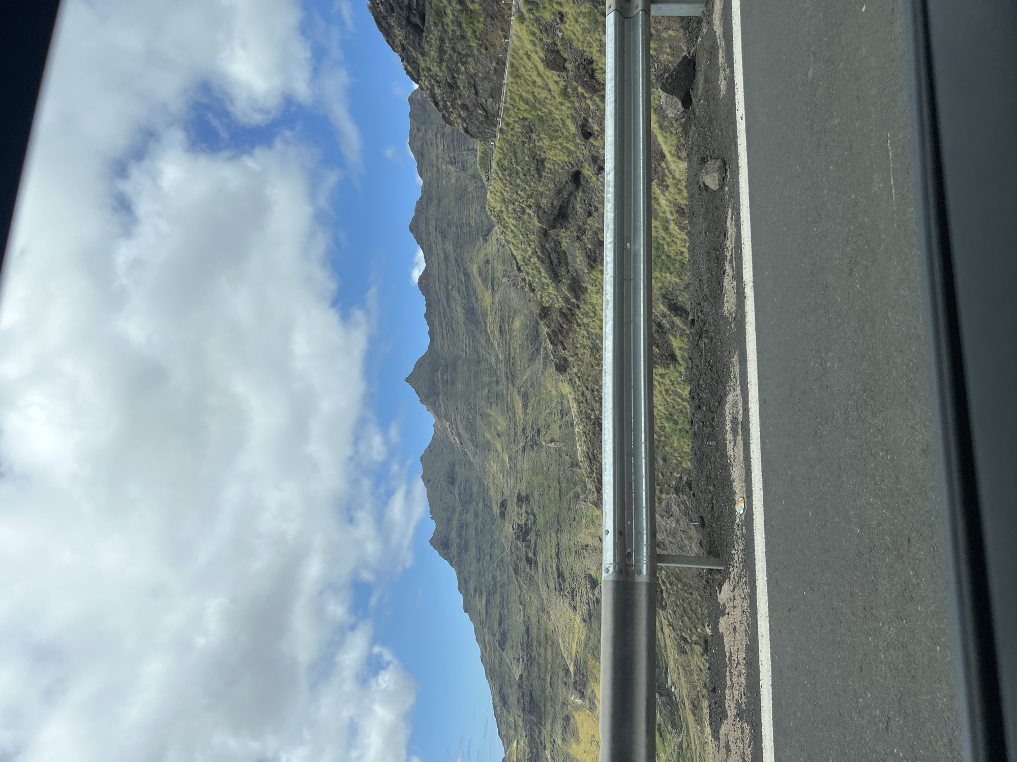A daytime, rotated view shows a landscape taken from a moving vehicle, specifically in Mogán, Spain. The sky features blue patches with white clouds. Adjacent to the sky, rugged, green-covered mountains rise sharply. A roadside barrier runs vertically through the image, separating the mountainous landscape from the road surface, which takes up the right side of the image. The view is partially framed by the vehicle's interior structure.