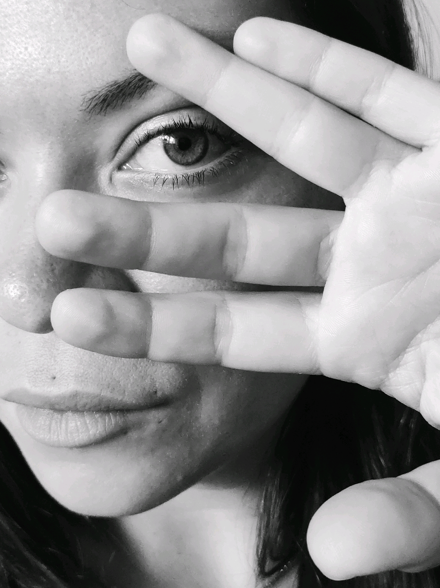 A black and white, close-up, portrait photograph captures a person's face. The subject is positioned in the center of the frame, with their right eye visible. Their hand is held up to their face, partially obscuring their features with their fingers layered across the lower half of their face and chin. The background is dark and indistinct, with strands of hair visible on the right side. The lighting highlights the texture of the skin and the details of the fingers and eye. The image does not contain any elements that would indicate its location in the city of Chaikovsky, Russia.