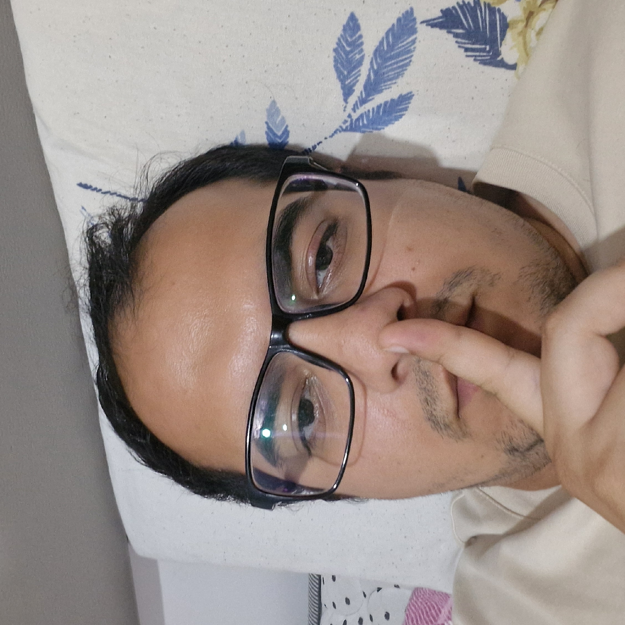 A man wearing glasses and a light-colored t-shirt is lying on a pillow with a floral pattern. His right index finger is held to his lips, signaling silence. The man's eyes are open and looking towards the camera. The image appears to be a selfie, taken from a close-up perspective. The background is a pillow with blue and white floral designs. The setting is indoors, possibly a bedroom. There is no information in the image that indicates the location is in Küçükçekmece, Turkey.