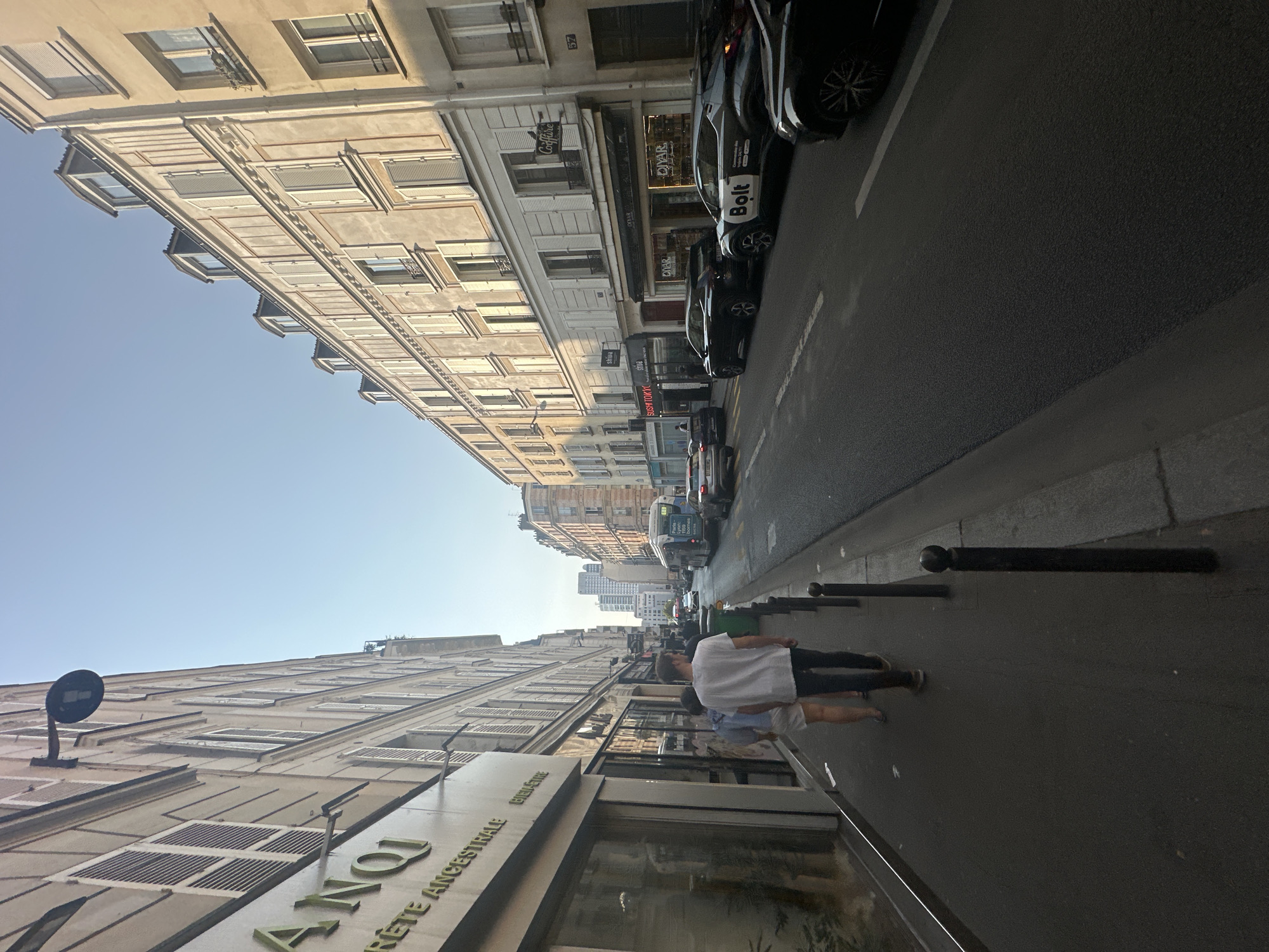 The image depicts a street scene in Paris, France, with typical Parisian architecture. On the left, a tall building with shuttered windows lines the sidewalk. A shop sign with the text "ANOQ" is visible. Two people, a man and a woman, are walking on the sidewalk away from the camera. The man is wearing a white t-shirt and black pants, and the woman is wearing a light-colored top and shorts.
The street itself is paved and appears to be a one-way street with cars parked and moving. Several vehicles are visible, including a white bus with "Paris-Lyon" text on it and a black car with "Bolt" branding. The buildings on the right side of the street are also tall and feature similar architectural elements like mansard roofs and shuttered windows. The sky is clear and a light blue, suggesting it is daytime, possibly late afternoon or early morning given the lighting. The scene conveys a sense of everyday urban life in a European city.