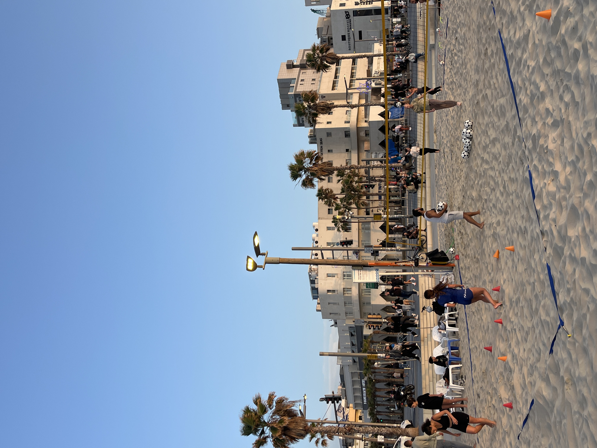 This outdoor image depicts a sandy beach area, likely in Tel Aviv, Israel, adjacent to a street lined with buildings and palm trees. The sky is clear and blue. People are engaging in beach activities, including soccer practice with multiple soccer balls placed on the sand. Cones are set up on the beach, possibly for training exercises. The beach area is bordered by a street with palm trees and various buildings, some identified as hotels. People are also sitting and standing along the street and near the beach. There is a beach volleyball net and a blue line in the sand.