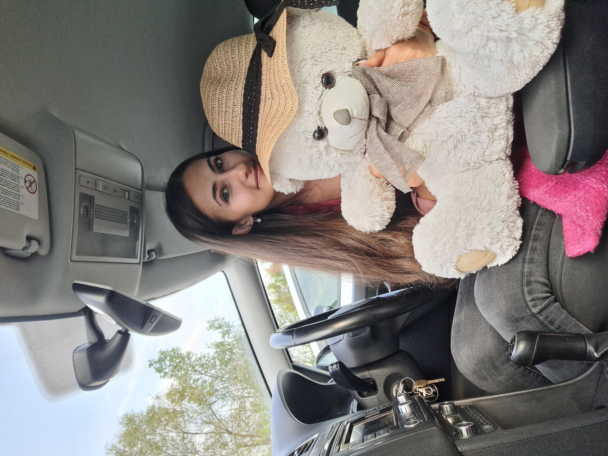 A young woman is pictured inside a car in Sofia, Bulgaria, holding a large white teddy bear. The image, originally rotated, shows the woman seated on the passenger side, smiling gently towards the camera. She has long, dark brown hair and is wearing a dark pink top and grey jeans. A pearl stud earring is visible in her left ear.
The prominent white teddy bear, almost as large as her upper torso, is adorned with a straw sun hat featuring a dark band and a beige and brown checkered bow tie. Its dark eyes and nose are clearly visible. The woman's hands are wrapped around the bear, suggesting a playful pose.
The car's interior is visible, featuring a grey headliner, a sun visor with a prominent "AIRBAG" warning label and safety diagrams, and a central overhead light console. A rearview mirror hangs from the windshield. To the left, a portion of the dashboard, a steering wheel, and a gear shift lever are visible, along with car keys in the ignition. The scene appears to be set during the daytime, with green trees and a bright blue sky visible through the car windows, indicating an outdoor location. A pink blanket or fabric is tucked into the seat beside the teddy bear. The overall mood conveyed is lighthearted and cheerful.
