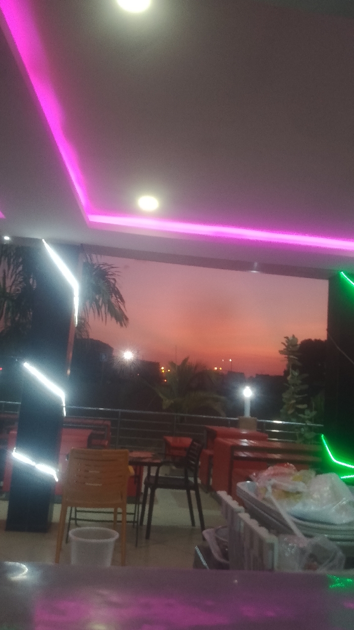 A restaurant or bar scene at sunset, showcasing a balcony view with outdoor seating. Orange and pink hues fill the sky beyond the balcony, contrasting with the interior's modern lighting. The ceiling has pink neon lights, and the interior features white and green light accents. Tables and chairs are visible, along with what appears to be a serving area or counter. Palm trees add a touch of tropical ambiance to the outdoor view.