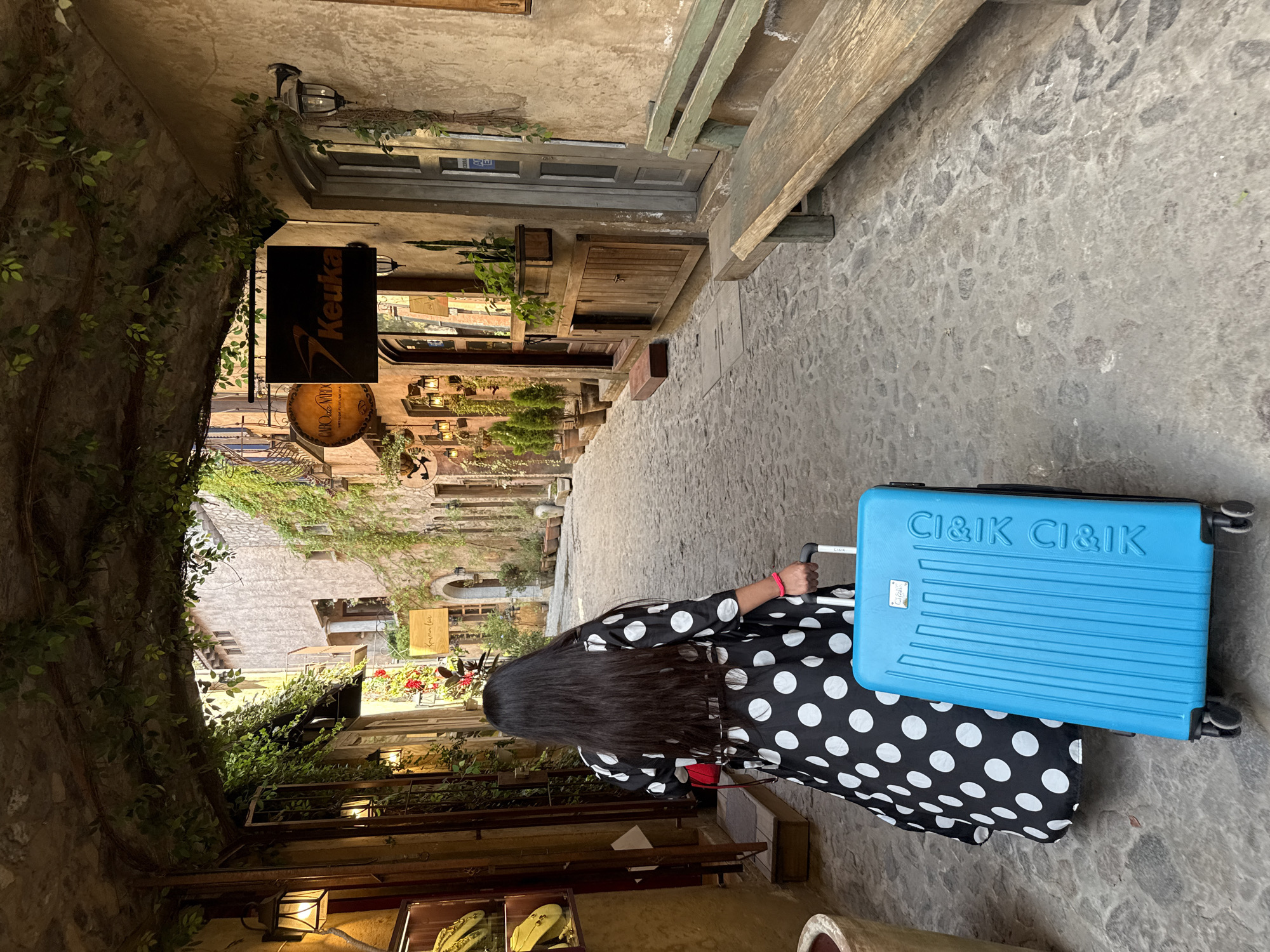 A woman with long, dark hair, wearing a black dress with white polka dots, pulls a blue suitcase down a narrow, stone-paved street. The street is flanked by old, stone buildings covered in vines and greenery. Signs for "Keuka" and "Riano de Ninix" are visible on the buildings. The image is possibly taken in Domingo Arenas, Mexico.