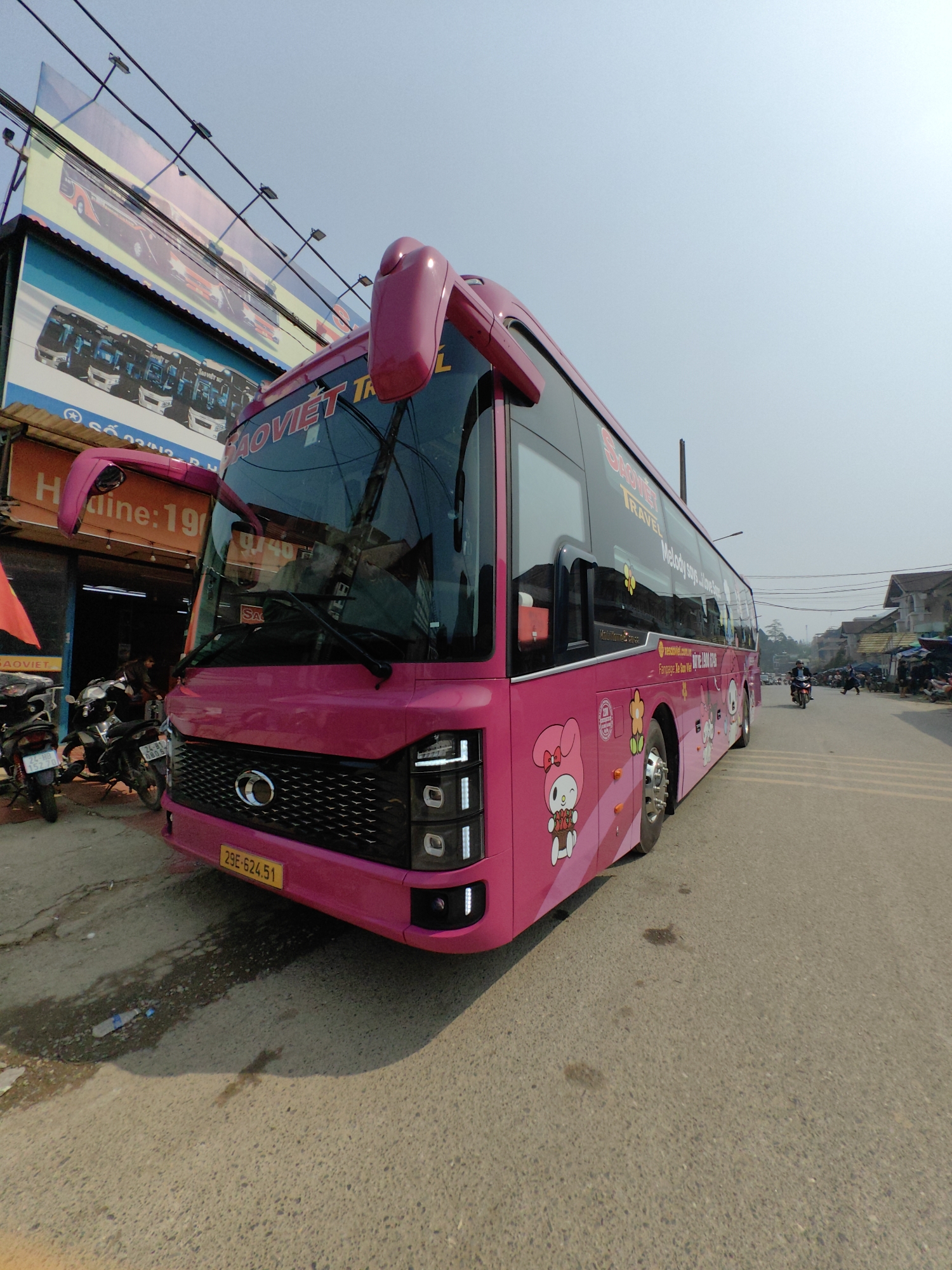 A bright pink passenger bus, branded "SAO VIET TRAVEL" with the license plate "29E-624.51" and "Melody says J'aime Sa Pa" text and Sanrio's Melody character decals, is parked on an asphalt road in Sa Pa, Vietnam. The bus occupies the left side of the frame, facing slightly right, with its front bumper positioned near the edge of a dirt/mud patch. To the left, a two-story commercial building with an orange-brown sign displaying "Hotline: 1900..." is visible. Above this building, a large billboard features images of similar buses against a light blue sky crisscrossed by overhead power lines. Several motorcycles are parked on the sidewalk near the building's entrance. Further down the street to the right, other buildings line the road, and a single individual rides a motorcycle away from the camera. The sky is clear and bright. A discarded clear plastic water bottle lies on the ground near a wet patch adjacent to the bus.