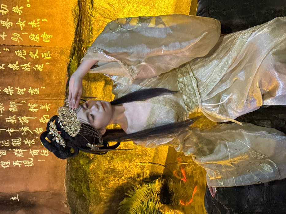 An East Asian female is positioned in what appears to be a cultural display environment within Yuzhong District, China. She is depicted from the waist up, facing slightly right with her head tilted down, her gaze directed towards the lower left. Her right hand is raised to her forehead, fingers touching her brow, in a gesture suggesting shielding her eyes or a contemplative pose.
She wears an elaborate traditional Chinese-style costume, primarily in shimmering golden fabric, resembling Hanfu. The garment features wide, flowing sleeves and intricate patterns on the fabric and a broad waistband. Her long, black hair is styled in an ornate updo, adorned with a large, golden headpiece or crown featuring intricate filigree, pearl-like accents, and a central red gemstone. Dangling golden chains extend from the headpiece to the sides of her face. Subtle makeup is visible, including defined eyebrows and eye makeup.
The background consists of a textured, reddish-brown wall illuminated by warm, golden light. Prominent Chinese calligraphy in gold characters is inscribed vertically across the upper portion of this wall. To the lower left, a dark, rocky pool of water is visible, containing several small, bright orange fish. Green foliage, possibly ferns, is also illuminated near the water feature. The woman's left hand rests on a dark, irregular, rock-like surface. The overall lighting is warm and artificial, creating a stylized atmosphere.