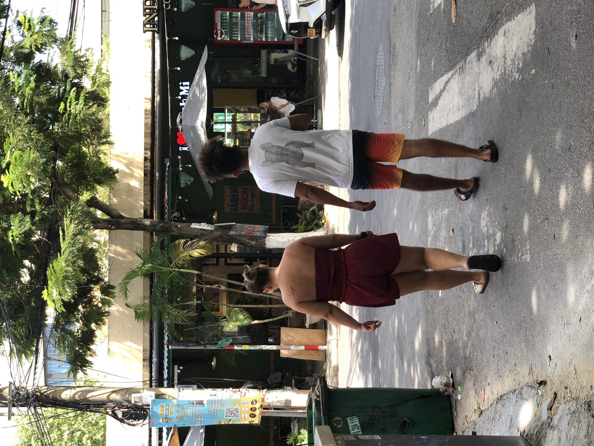This media file shows a street scene in Đà Nẵng, Vietnam. Two individuals are walking on a paved street, with buildings and lush greenery on one side. The person in front, viewed from the back, is wearing a white t-shirt with a graphic print and ombre shorts, with curly hair. The person behind, also viewed from the back, is wearing a dark red skirt and has their hair in a bun; a small tattoo is visible on their upper back.
The scene appears to be during the day, with sunlight casting shadows on the ground. Several signs are visible, including one for "BANH MI NGON Gia truyền" (Delicious Baguette, traditional recipe) and another advertising a "DANANG CRAFT BEER FESTIVAL" with dates and activities. Other text includes "IND," "CC," and "MATCHA LATTE," suggesting a commercial area with food and drink establishments. A Coca-Cola refrigerator is visible in one of the buildings. There are also a number of electrical wires and a green trash bin in the foreground. The overall impression is of a bustling urban environment, possibly a commercial street or sidewalk in a Vietnamese city. The people are walking casually, suggesting a relaxed atmosphere.