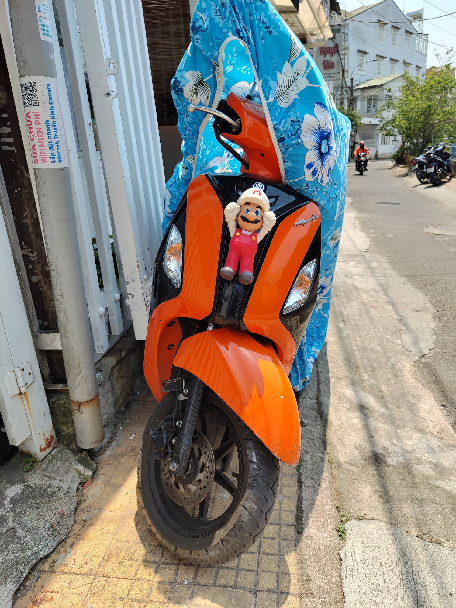 An orange and black scooter with a Mario figurine on its front is parked on a sidewalk in Thành phố Đà Lạt, Vietnam. The scooter is partially covered with a blue floral-patterned sheet. Next to the scooter is a white metal fence with a sticker affixed to one of the posts. The sticker contains Vietnamese writing. In the background, there is a road with other parked scooters and a building with the words "Nguyet Vien" visible. A person on a scooter is driving down the road in the background. The day is sunny, and the street is relatively quiet.