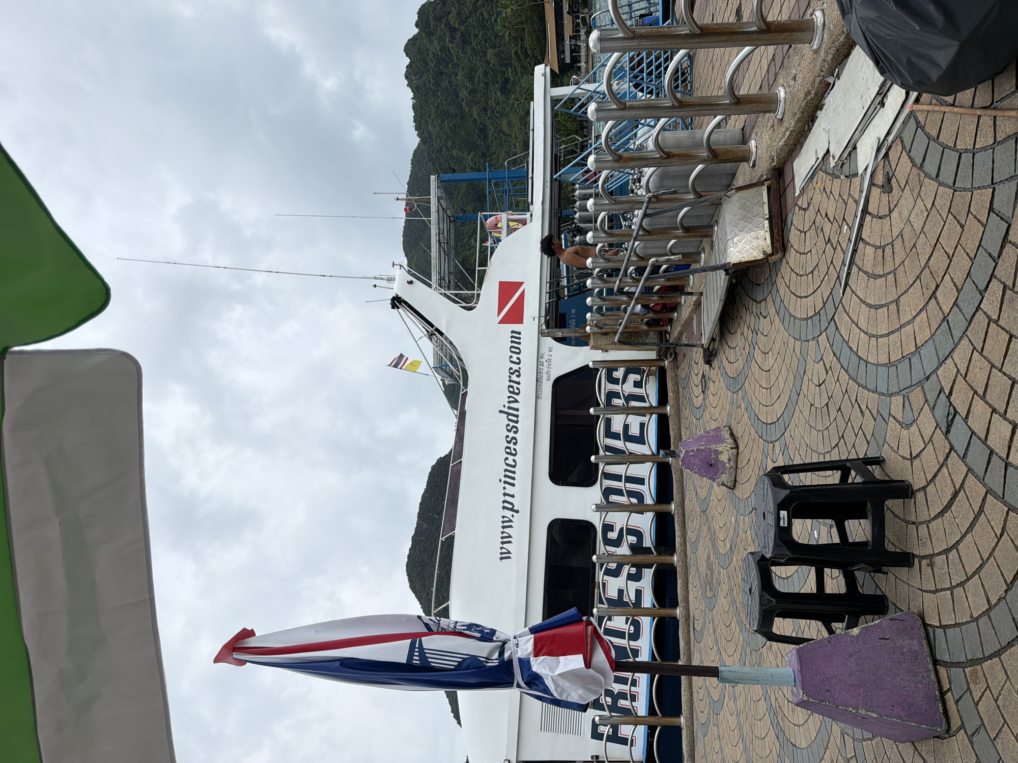 In Ao Nang, Thailand, a large white dive boat named "PRINCESS DIVERS" is docked at a pier under an overcast sky. The boat has multiple windows and the website "www.princessdivers.com" is visible on its side. A Thai flag flies from the boat's mast. In the foreground, a colorful umbrella is folded and tied to a pole, adjacent to two black plastic stools. A ramp leads from the pier onto the boat, and several dive tanks are lined up along the railing. A man, shirtless, stands near the ramp, appearing to be working or waiting. The pier is paved with a circular pattern of brown and grey tiles. In the background, a lush green, mountainous island rises from the sea.
