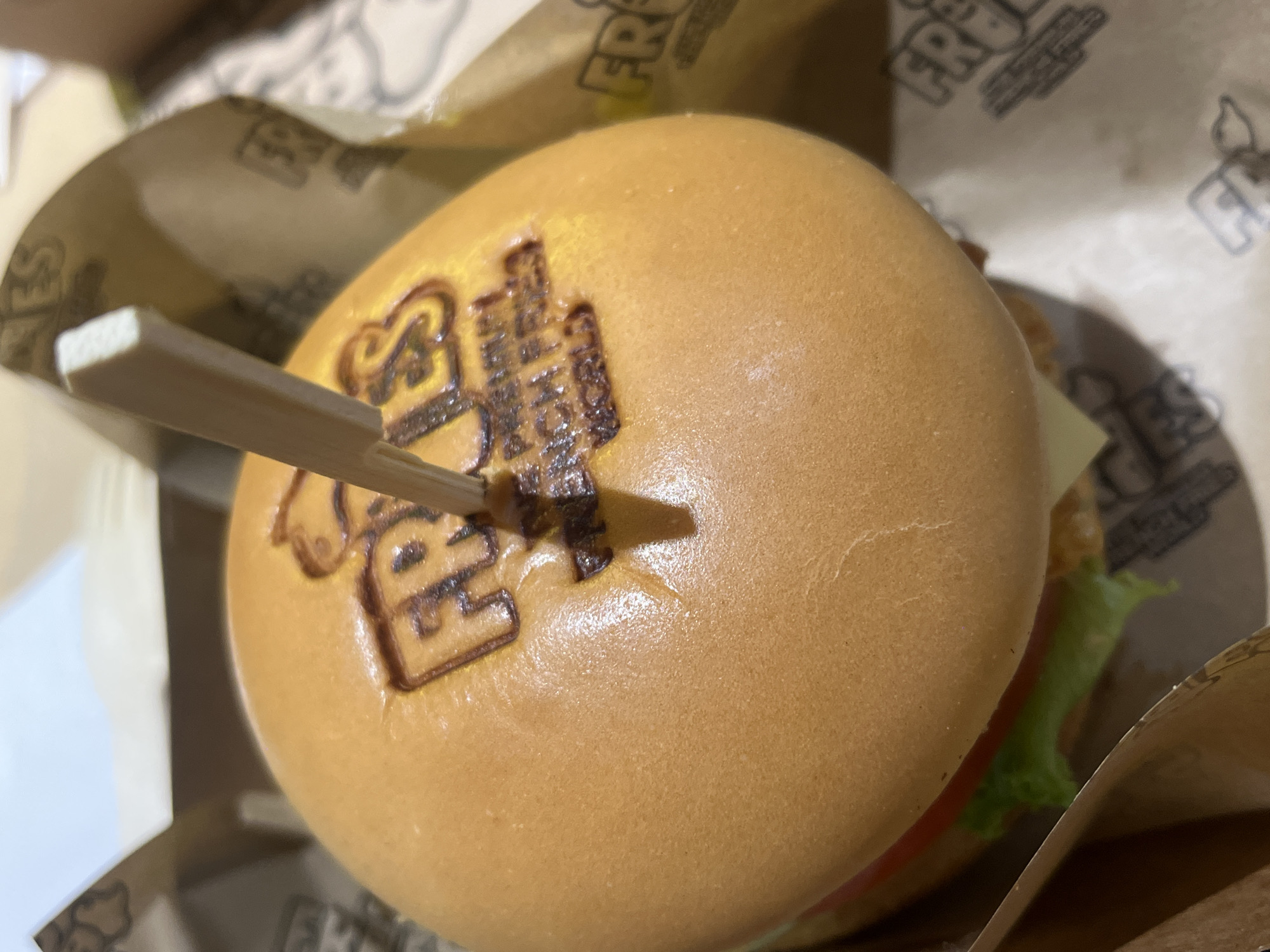 A close-up shot of a burger, secured with a toothpick, resting inside a takeout container. The burger bun is branded with lettering. The container displays printed text and logos. Components of the burger's filling such as lettuce and tomato are visible. The scene is situated in Thủ Đức, Vietnam, as indicated by the "FRJ's" branding.