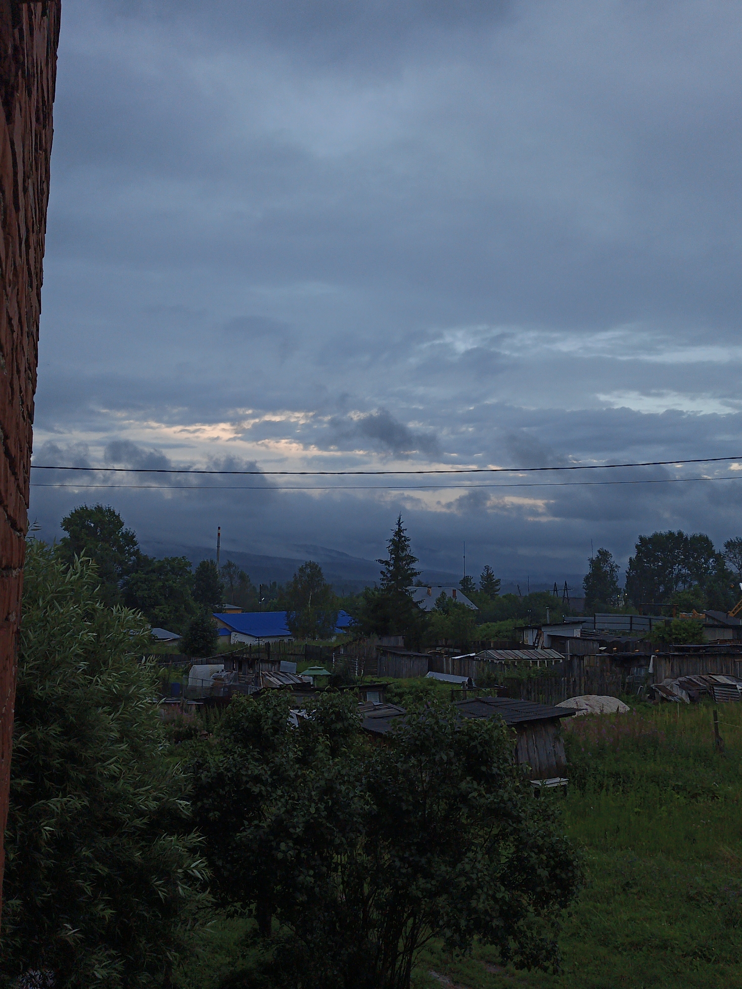 The image depicts a rural or suburban landscape under a cloudy sky. In the foreground, on the left side, a portion of a brick wall is visible, suggesting the viewpoint is from inside or near a building. The middle ground reveals a collection of structures, including houses with blue roofs and various wooden sheds and fences. Trees and vegetation are abundant throughout the scene, with dense foliage in the foreground and scattered trees and bushes behind the buildings. In the distance, a hazy mountain range or hills can be seen through a layer of fog or low clouds. A tall, slender structure, possibly a chimney or a radio tower, rises above the trees in the mid-distance. Two horizontal wires are stretched across the upper part of the scene. The overall atmosphere is overcast and subdued, hinting at either dawn, dusk, or impending bad weather. The scene is consistent with a residential area in a rural or semi-rural setting in Medvedyovka, Russia. No people or animals are visible in the frame.