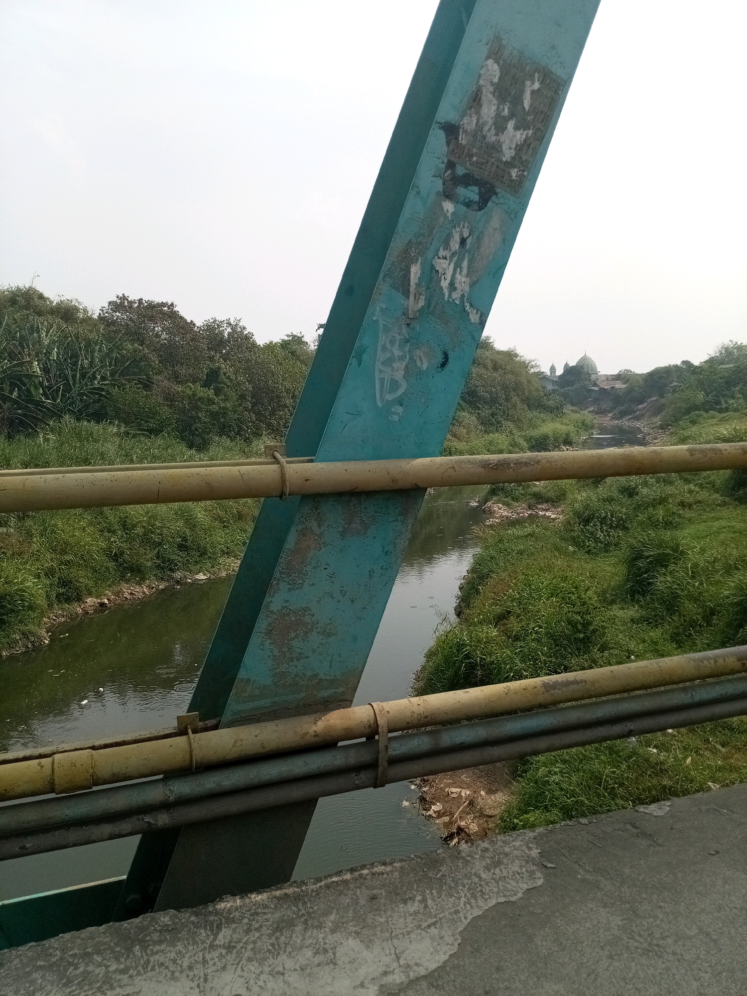 The image shows a view from a bridge in Karangsatria, Indonesia. A greenish-blue steel beam, part of the bridge structure, is prominently in the foreground, angled from the top left towards the center bottom. Attached to this beam are two horizontal pipes, one yellow and one gray, running across the frame. Below these pipes, a narrow river flows, bordered by lush green grass and vegetation on both banks. In the background, beyond the river and trees, are two dome-shaped structures, possibly part of a mosque or other religious building. The sky is overcast and light gray. There are no people visible in the image. The scene depicts an outdoor environment with man-made structures (bridge components, pipes) and natural elements (river, vegetation, sky).