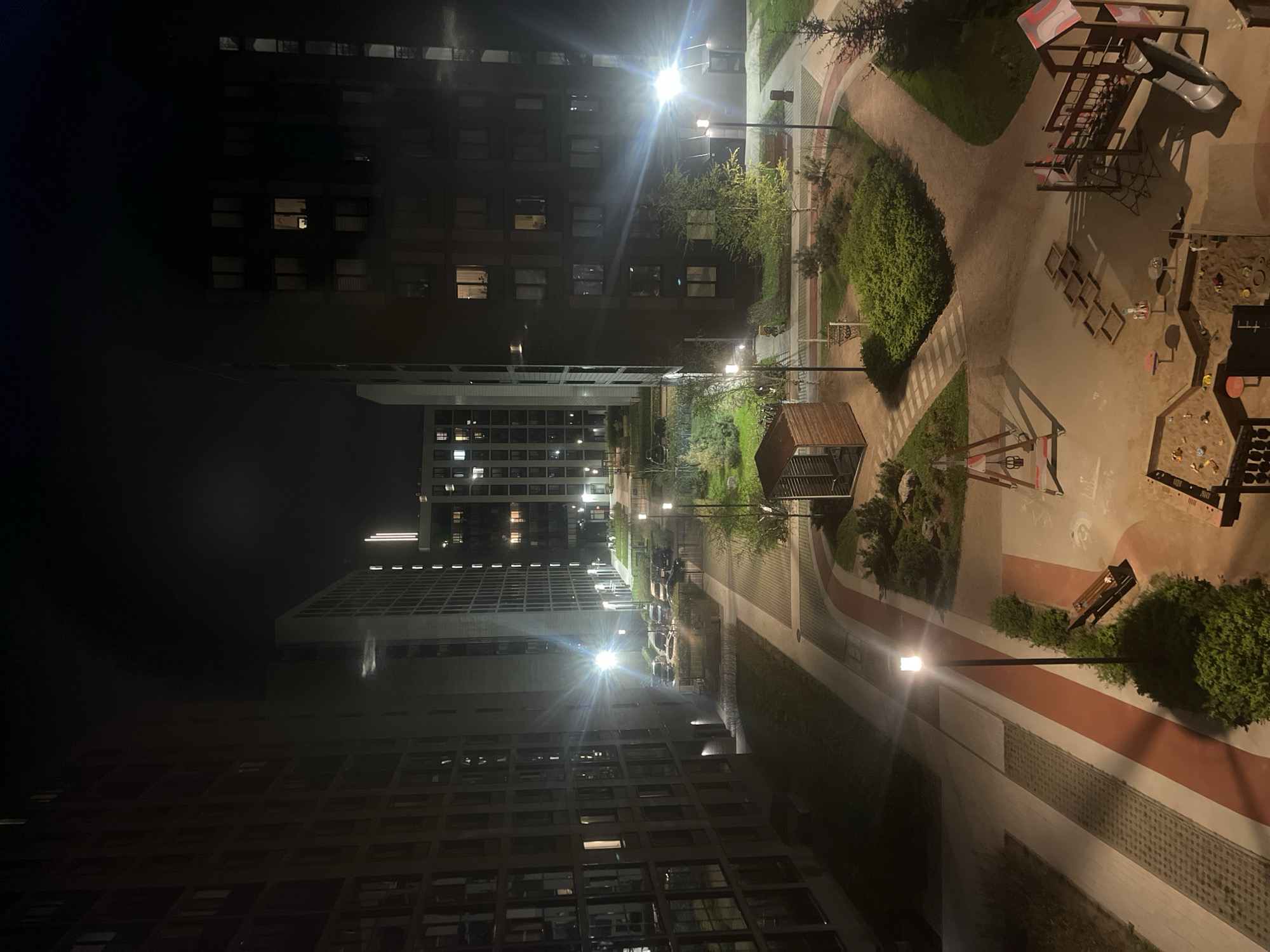 The image shows a nighttime view of a courtyard located in what appears to be a residential area in Saint Petersburg, Russia. High-rise apartment buildings surround a central recreational space. The courtyard contains a children's playground with structures like slides and sandboxes. Several paths are visible, illuminated by streetlights, creating a network of walkways through the landscape. Vegetation, including trees and bushes, are integrated into the courtyard's design. The lighting suggests a quiet urban atmosphere, typical of a residential neighborhood at night. There are no people visible in the frame.