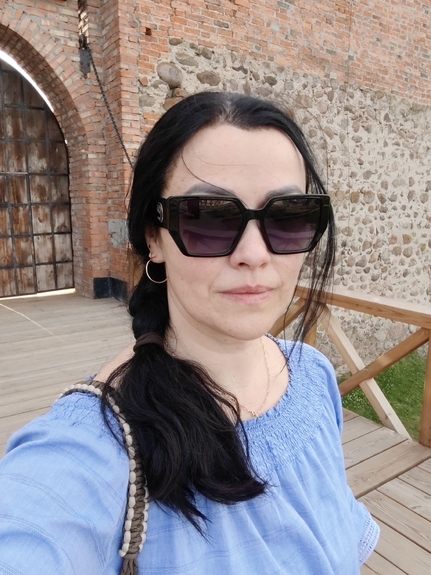A woman is taking a selfie while standing on a wooden boardwalk in front of an old brick and stone wall. She has long, dark hair styled in a braid and is wearing large, dark sunglasses and a light blue off-the-shoulder top. A woven bag strap is visible across her shoulder. Behind her, a large arched wooden gate is visible within the brick structure, suggesting a historical or castle-like setting. The wall is made of rough-hewn stones interspersed with bricks.