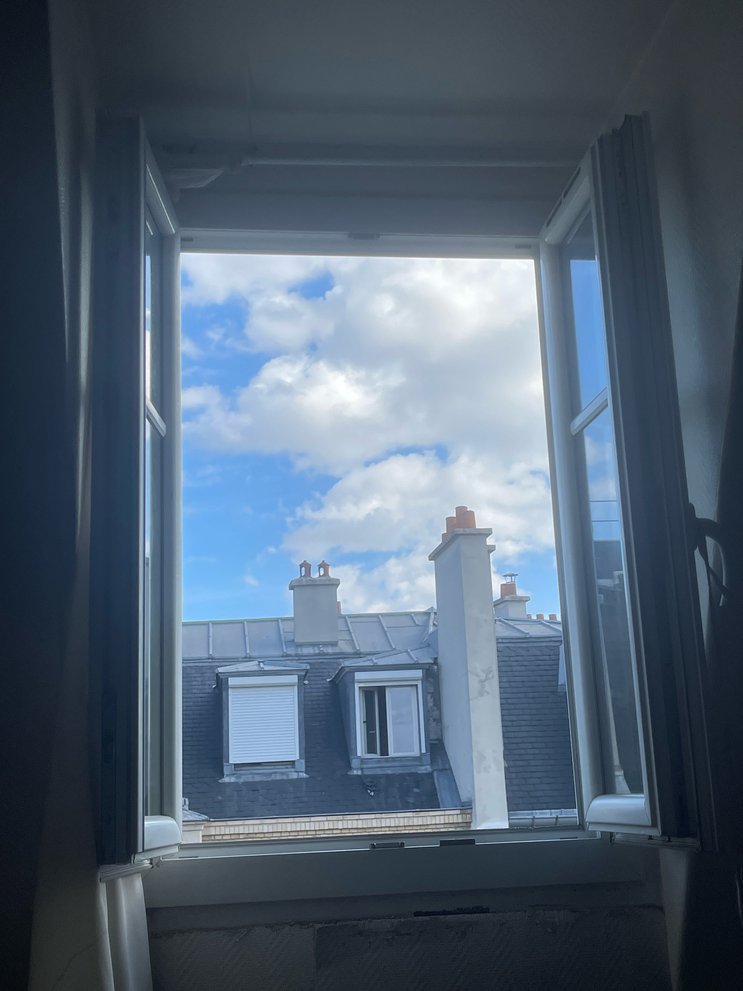 The image shows a view looking out of an open window in what appears to be an apartment building in Paris, France. The window frames, made of white material, are open inwards and outwards. Outside the window, the sky is blue with scattered white clouds. Below the sky are the grey, slate roofs of Parisian buildings, characterized by their mansard design. Several chimneys, typical of Parisian architecture, are visible on the rooftops. One chimney has two distinct openings, while another has three. One of the buildings has a white roller shutter partially closed over a window. The interior of the room from which the photograph is taken is dark, with curtains visible on the sides of the window. The bottom of the window frame shows a textured surface, possibly wallpaper. The scene is static, with no people or animals present. The overall impression is that of a typical urban residential view.