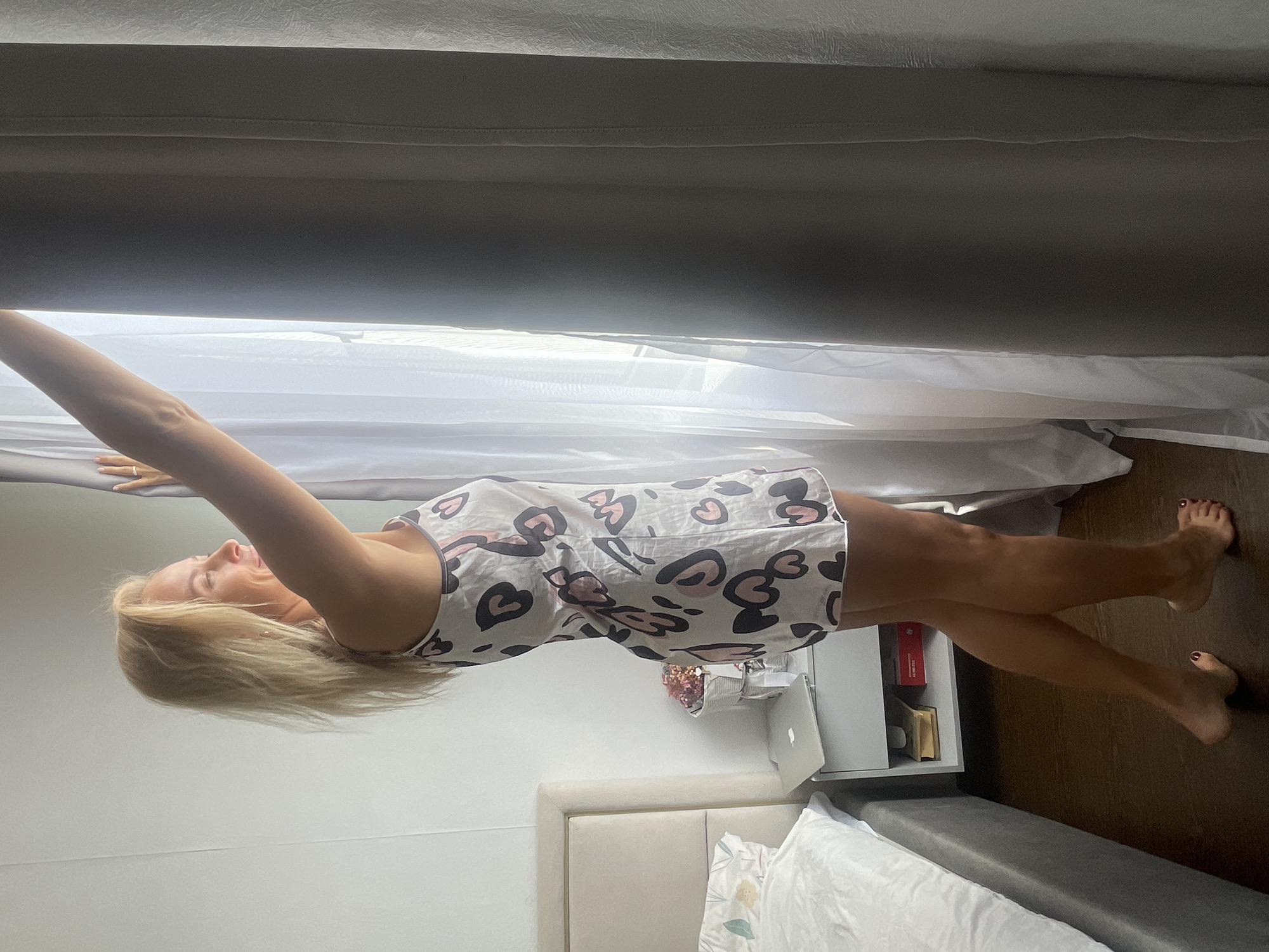 A medium-shot, indoor photograph presents a woman in a printed, white pajama set, possibly cotton, decorated with black and pink heart shapes. She is standing upright, arms extended overhead, one hand appearing to touch the top of a curtain or shade. The woman has fair skin and blonde hair.
The setting includes a bed with a light-colored headboard, white bedding, and a grey bed frame. A small white bedside table is visible, holding a laptop, a vase with flowers, and books. The flooring appears to be dark wood. A closed curtain or shade occupies the upper portion of the frame. The location is not verifiable as Baranovka, Russia.