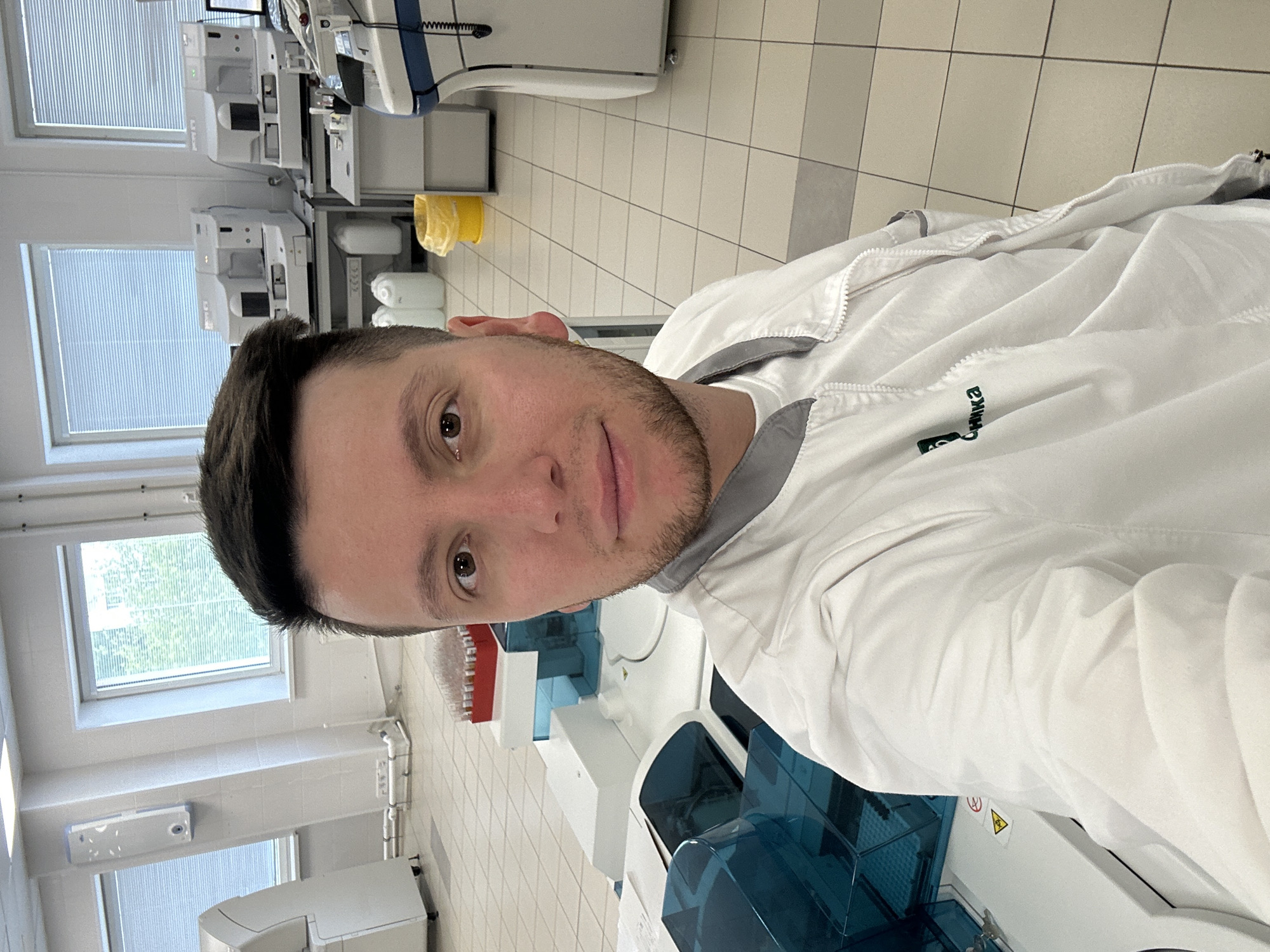 A medium shot depicts a man indoors, seemingly in a laboratory setting. The man has short, dark hair, light skin, and brown eyes. He sports a neatly trimmed beard. He is wearing a white lab coat with a gray trim around the collar and zipper.
The background reveals a tiled floor and white-walled room. Visible through a window are green trees. Scientific equipment, including testing machines with blue and white components, are in the scene. Another machine is against the back wall.
The location is likely in Moscow, Russia, due to the Cyrillic letters on the lab coat.