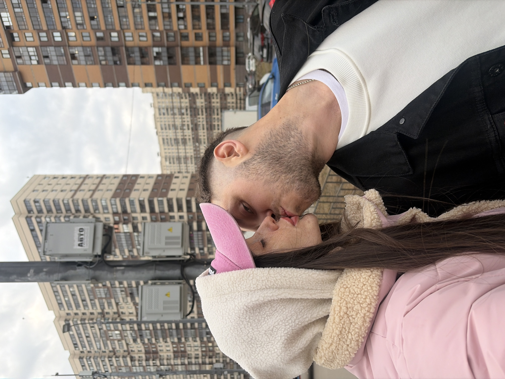 A man and a woman are kissing in front of high-rise apartment buildings in Saint Petersburg, Russia. The man has short dark hair and a beard, wearing a white shirt with a black jacket. The woman has long dark hair, wearing a light pink jacket with a white furry hood and pink bunny ear hat. The photograph appears to have been taken from a low angle, with a light grey sky. There is a metallic pole with grey electrical utility boxes.