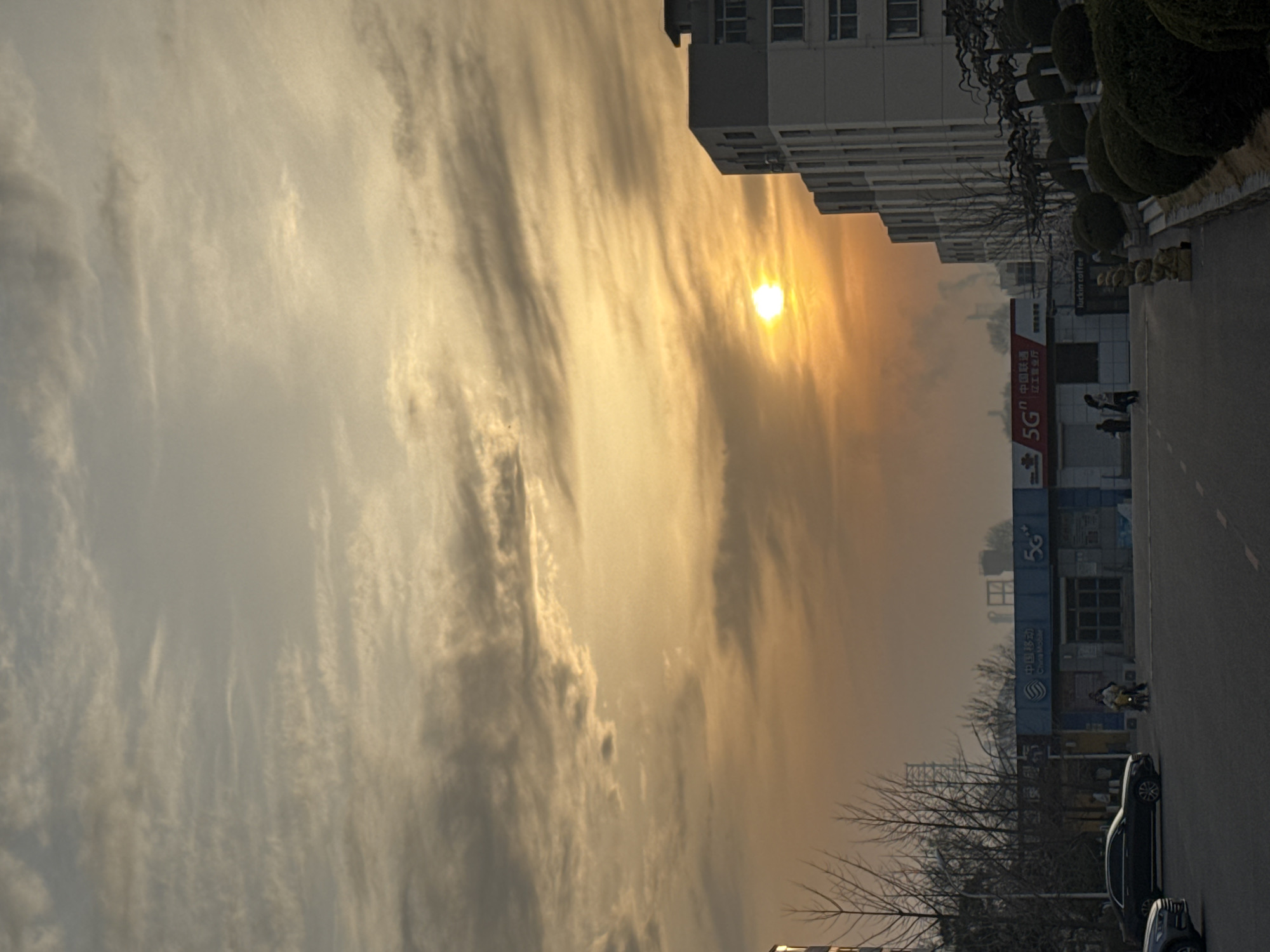 The image shows a cityscape in Zhongtun, China, under a sky with thin, streaky clouds illuminated by a bright sun. Along a street are parked cars and a building with a "5G" logo. Some people are walking on the sidewalk. Trees are bare of leaves.