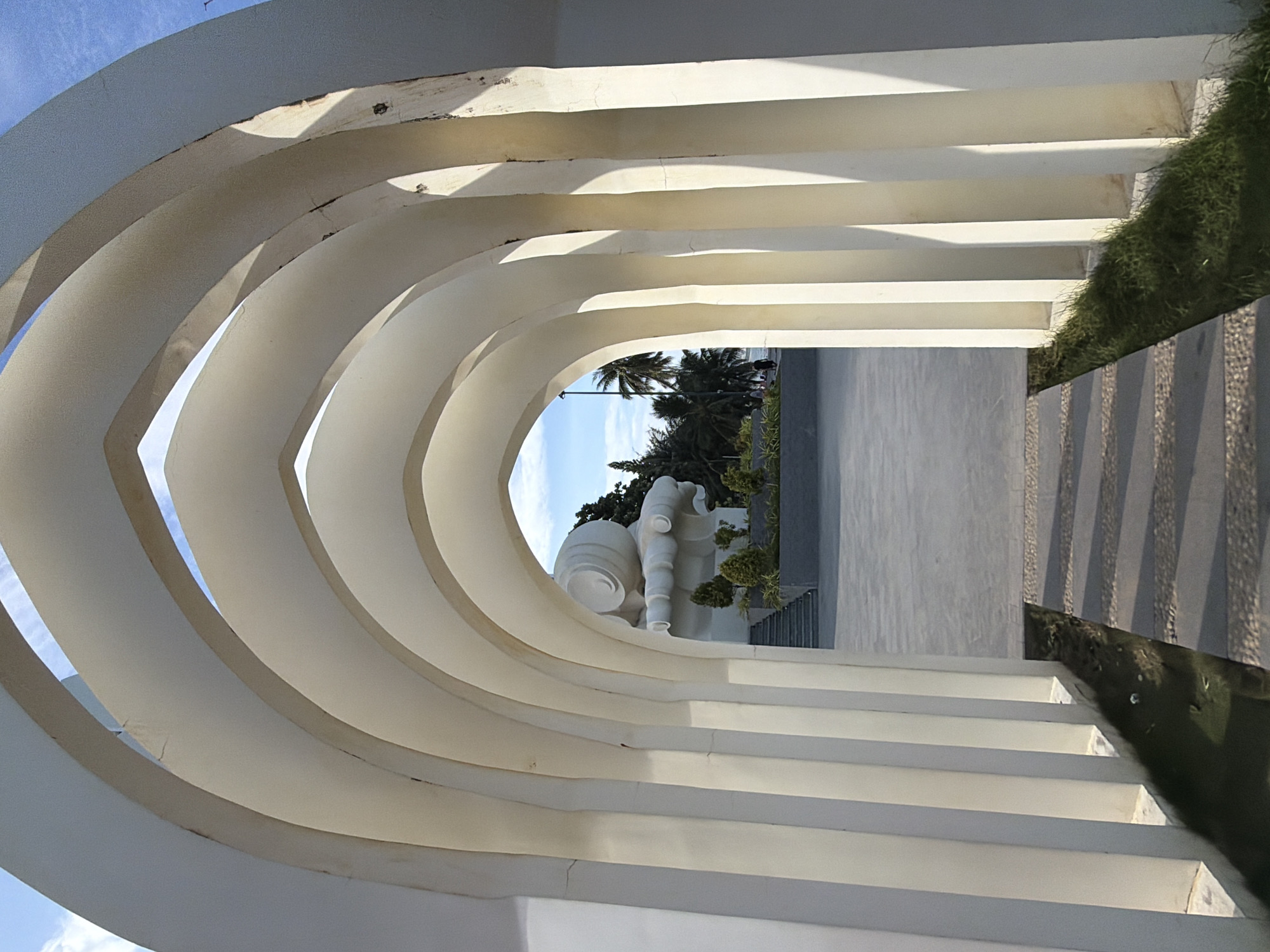 The image shows a view through a series of arched white structures, resembling a portico or arcade. Beyond the arches, a large, white, abstract sculpture is visible, resting on a raised platform. Steps lead up to this platform. The sculpture appears to be lying down, with rounded, curved forms. In the background, there are palm trees and a bright blue sky with some clouds. To the right of the sculpture, a hint of the ocean or a body of water can be seen. The ground in front of the arches is paved with concrete and stone, with a narrow strip of green grass bordering one side. There are no people clearly visible in the foreground. The overall impression is of a modern architectural setting with a prominent art installation. This scene is located at the Tran Phu Bridge in Nha Trang, Vietnam, specifically at the gateway to the bridge which features this sculpture and surrounding plaza.