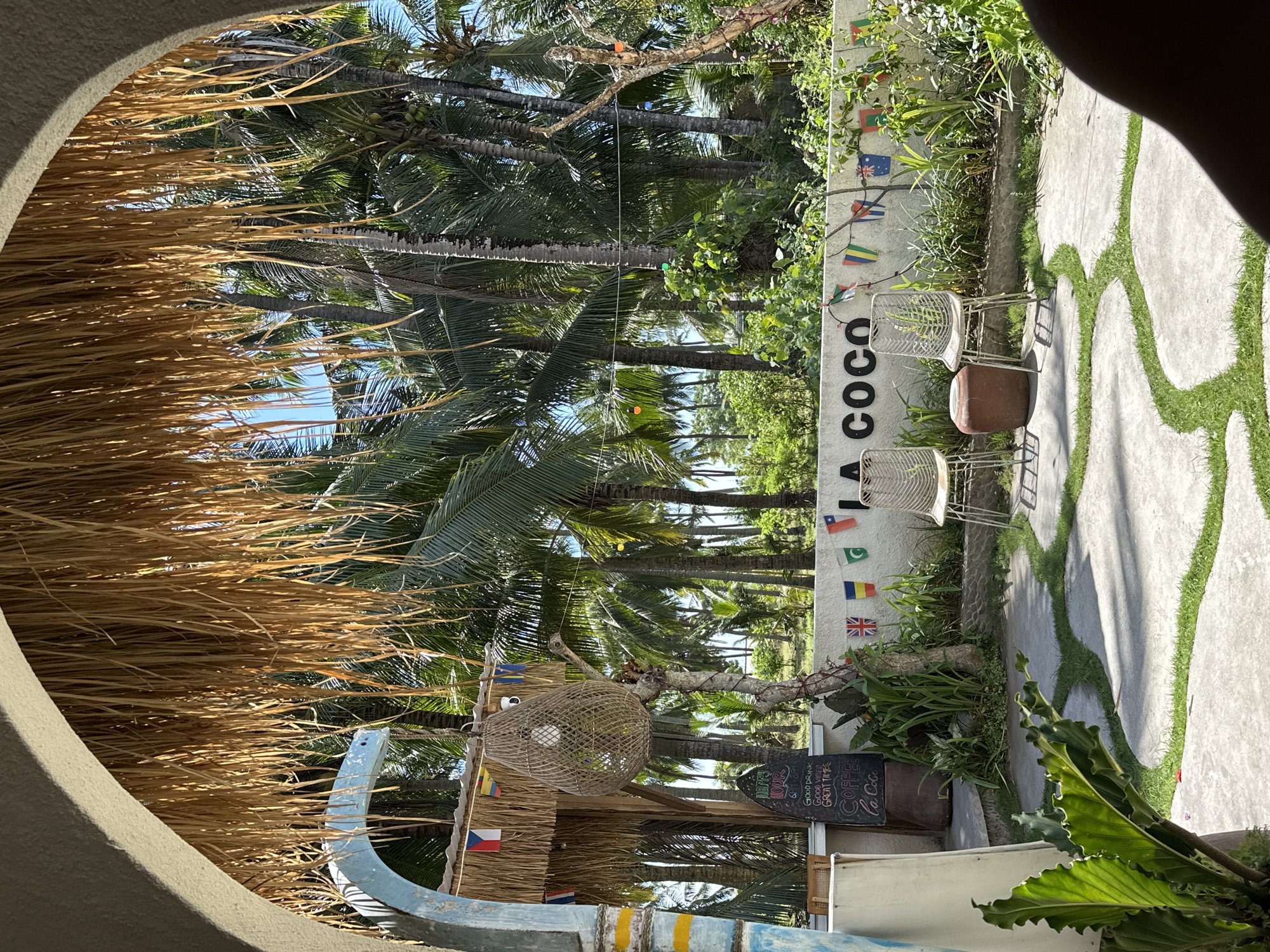 The image shows an outdoor scene, possibly a café or restaurant, framed by a circular opening or window. The scene includes tall palm trees, tropical foliage, and a white wall adorned with the name "La Coco" vertically printed in black letters. The wall also features a string of flags representing various nations.
In front of the wall, there are two metal chairs and what appears to be a dark-colored container or bin. A chalkboard menu with the words "Happy Hour," "Good Drink, Good Vibes, Great Times," and "Coffee la Coco" is visible near the bottom of the image.
The foreground features part of a thatched roof structure and a section of a blue and yellow boat or canoe-like object. A woven hanging lamp is visible amidst the foliage and structures. A path with green grass in between the concrete stones is also visible in the background.
Based on the "La Coco" name and tropical setting, the location is likely in Nusa Penida, Indonesia.