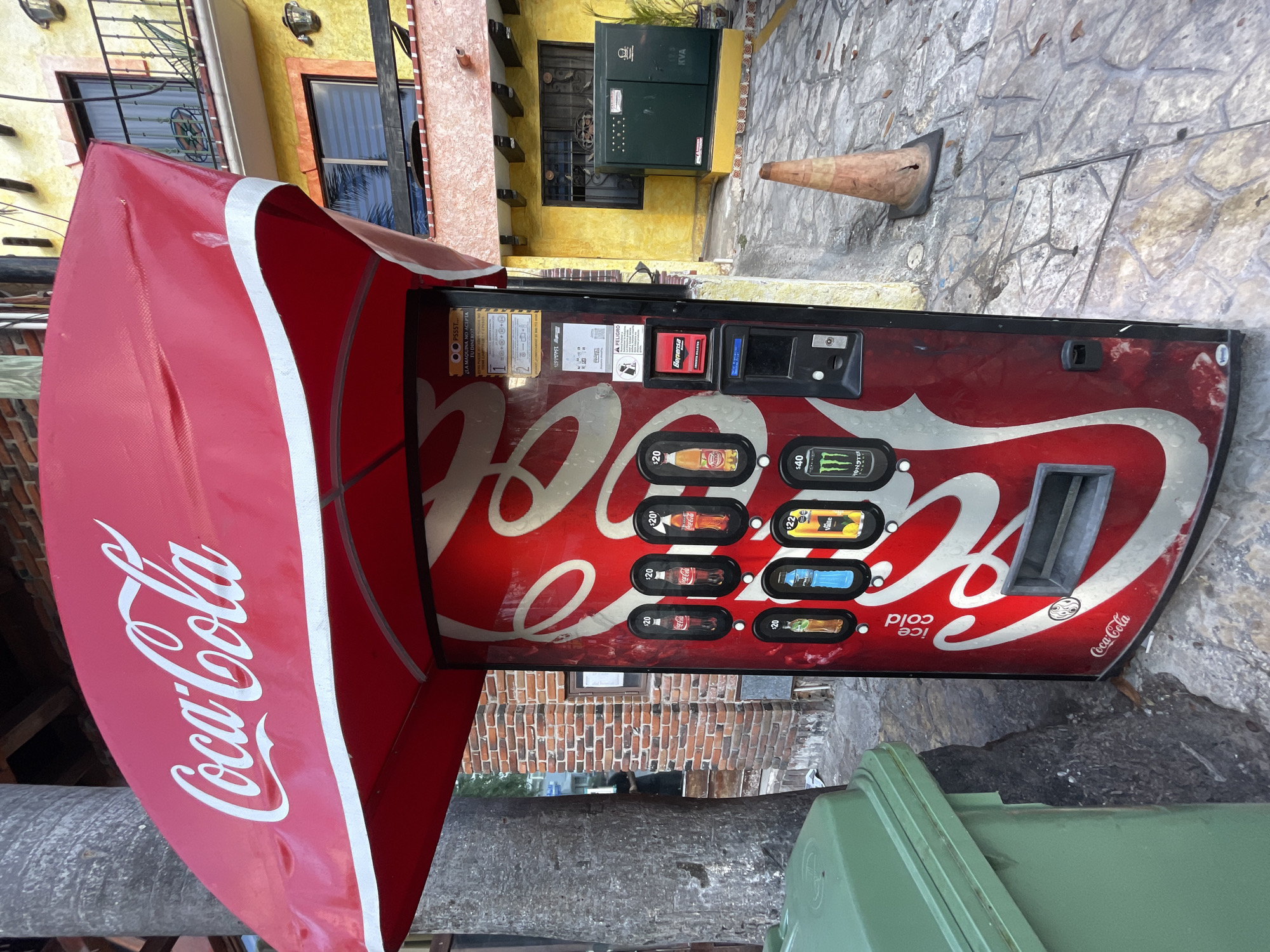 The image shows a weathered Coca-Cola vending machine in Playa del Carmen, Mexico. The machine is red and features the Coca-Cola logo prominently, with prices for beverages ranging from $20 to $40. A red Coca-Cola branded awning extends over the machine. The machine is positioned on a stone-paved area, next to a green waste container. Behind the machine, there is a yellow building, a brick wall and a cone.