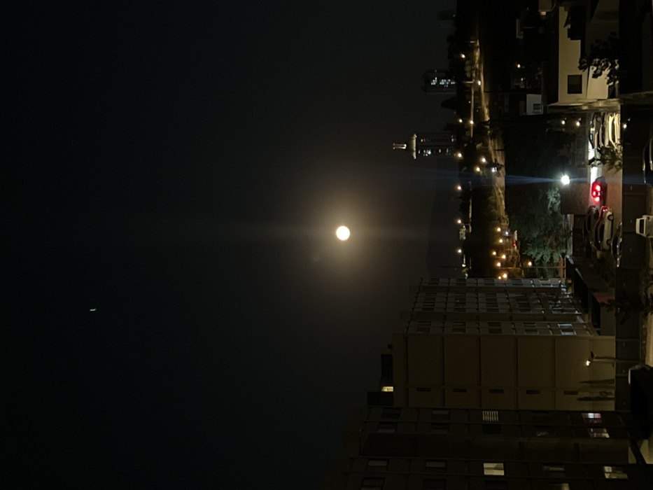 The image depicts a nighttime urban landscape in León, Guanajuato, Mexico, captured from an elevated perspective. The sky is dark, featuring a bright, full moon positioned centrally, emitting a distinct vertical light ray. A faint, crescent-shaped object is visible in the upper left quadrant of the sky.
In the left foreground, a multi-story, light-colored residential building occupies a significant portion of the frame, with several windows displaying interior illumination. Below this building, a ground-level parking area is visible, containing multiple parked vehicles; one car's red taillights are illuminated.
The midground reveals a city sprawl characterized by numerous streetlights and illuminated buildings. A prominent, tall building with a distinctive illuminated crown, identified as the Poliforum León Tower, stands slightly to the right of the image center. Other smaller buildings and patches of vegetation are also visible. Distant hills or mountains are faintly discernible on the horizon line. No people are visible.