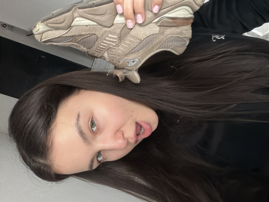 A young adult female is depicted in a close-up, chest-up portrait within an indoor setting. She possesses long, dark brown hair, fair skin, and light-colored eyes. Her mouth is open in an 'O' shape, and she gazes directly at the camera. She is wearing a dark, long-sleeved top with a small, reflective '7' emblem positioned on the left side of her chest. Her right hand, exhibiting pink-painted fingernails, holds a single light brown/beige athletic shoe, specifically a bulky sneaker, placed near her right ear and shoulder. The background consists of indistinct light-colored walls or surfaces, with a dark, rectangular object visible behind her head on the right side, above which a partial '6' character is faintly discernible. The specific location within the city of Sofia, Bulgaria, cannot be determined from the visual evidence provided.