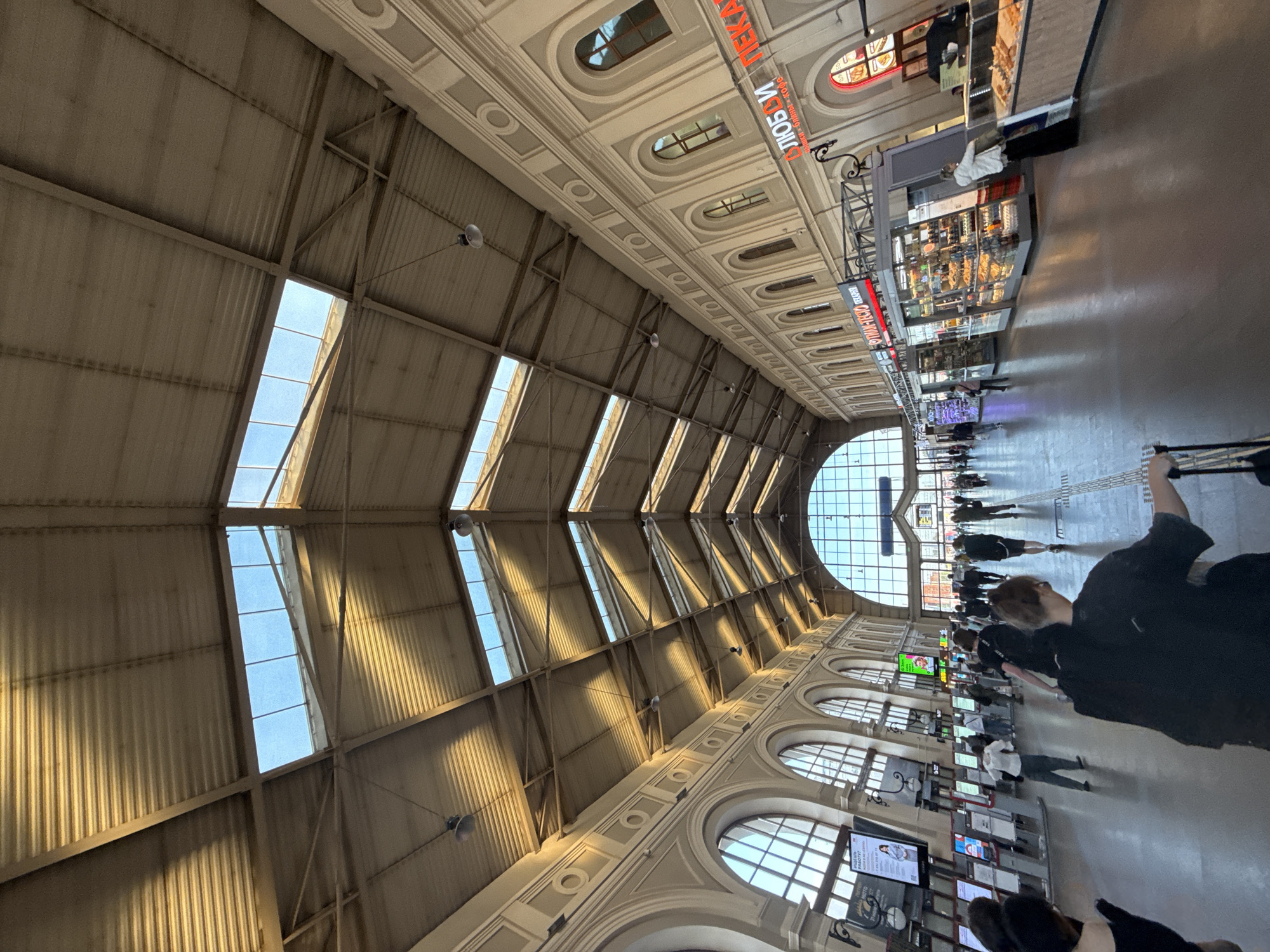 The image depicts the vast, high-ceilinged interior of a large public building, likely a train station or a grand concourse. The perspective is an upward-angled shot along the length of the hall.
The ceiling features an industrial design with corrugated metal panels supported by a network of metal beams. Several long, rectangular skylights are integrated into the roof, allowing bright natural light to illuminate the space and cast distinct shadows. Below the roof structure, the walls are adorned with ornate classical architecture, including decorative moldings, recessed panels, and arched windows, some of which are visible on the right.
Along the right side of the hall, a series of commercial kiosks or stalls are set up, offering various goods, including food items. A prominent red sign above one of these stalls displays Cyrillic text: "DИKОBЛИ ПЕКАР..." with smaller text beneath reading "домашняя выпечка и кофе" (homemade pastries and coffee). Another partially visible red sign begins with "ФРАНЦУЗСКАЯ..." (French...).
Numerous people are present on the polished floor, walking and standing. Some individuals appear to be browsing the stalls, while others are simply moving through the area. In the foreground on the right, a person in a black shirt is partly visible, holding the handle of what appears to be a wheeled suitcase. At the far end of the hall, a large, semicircular arched window with a grid pattern allows more light into the space. The bright light suggests it is daytime with clear weather. Speakers are mounted from the ceiling beams. The scene portrays a bustling, functional public environment.