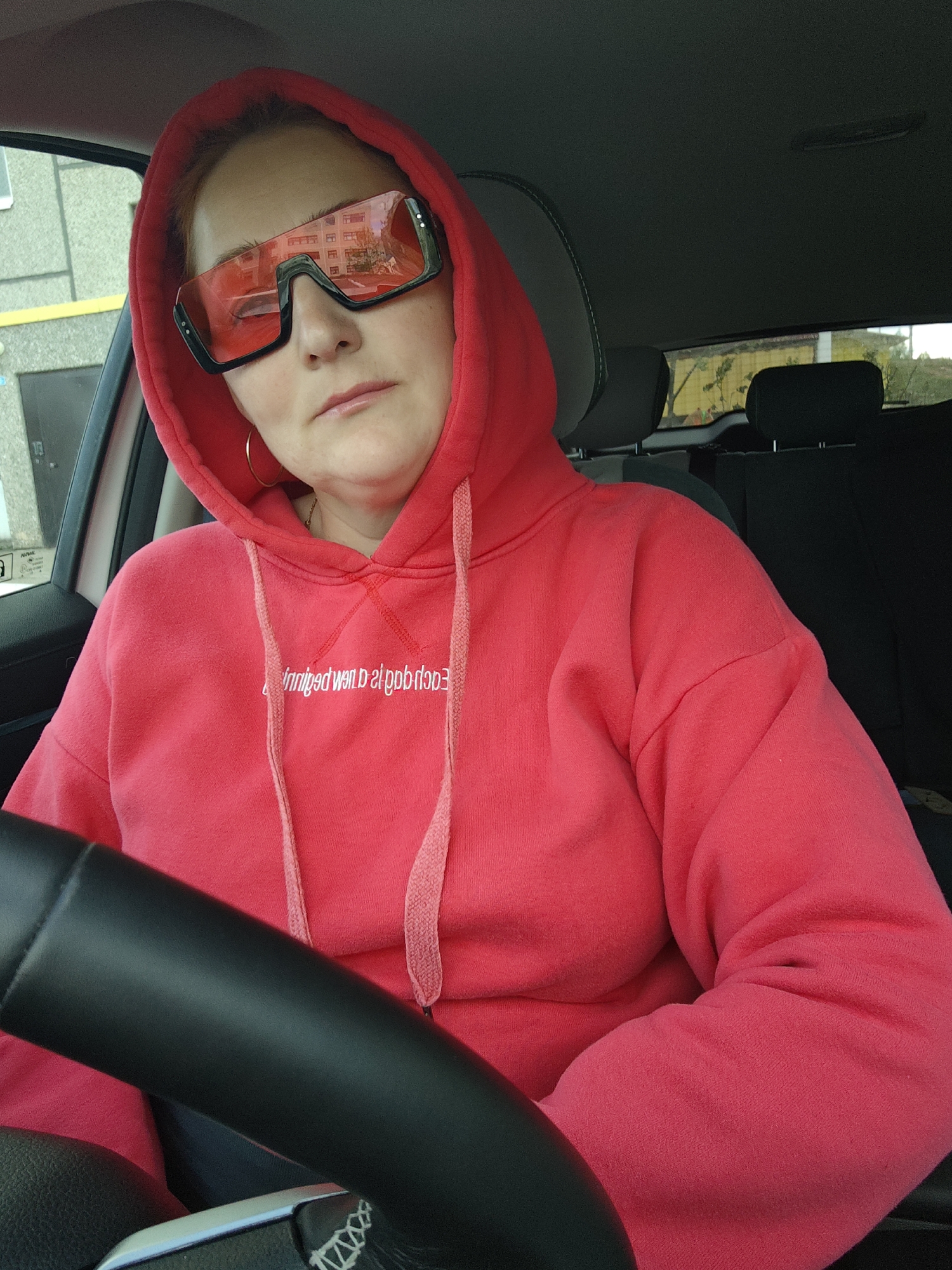 The image depicts a fair-skinned woman in the driver's seat of a car. She is wearing a red hoodie with the hood up, and large, futuristic-looking sunglasses with red lenses and a black frame. Her facial expression is neutral, and her gaze is directed slightly downward. She is wearing gold hoop earrings.
The woman is positioned behind a black steering wheel. The interior of the car is visible, with dark gray seats. The scene is taken from inside the car looking outward; buildings are visible through the windows. Based on the exterior architecture, window reflections, and surrounding buildings, the location is likely Markovskii, Russia.