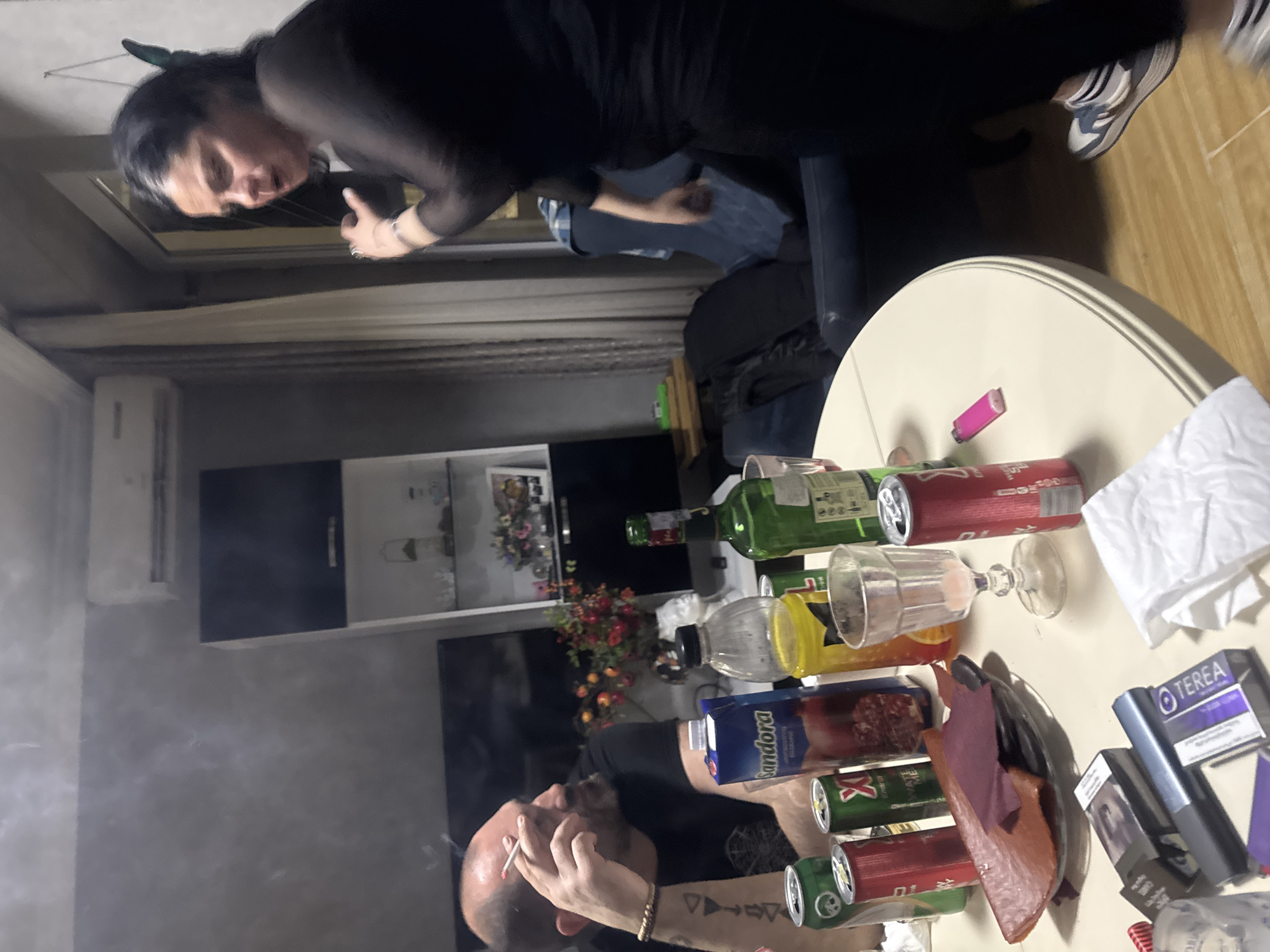 Two people are present in a dimly lit room. One individual, dressed in black, appears to be leaning over a round table. The second person, visible from the torso up, is seated near the table, smoking a cigarette. The table is cluttered with bottles, cans, glasses, cigarette packs, and what appears to be a plate with food. The background shows a cabinet with decorative items, suggesting an interior setting. The scene exudes a casual, indoor gathering atmosphere. While there is no definitive indication in the image that this location is specifically in the city of Tbilisi, Georgia, it is implied that the image was taken in this area.