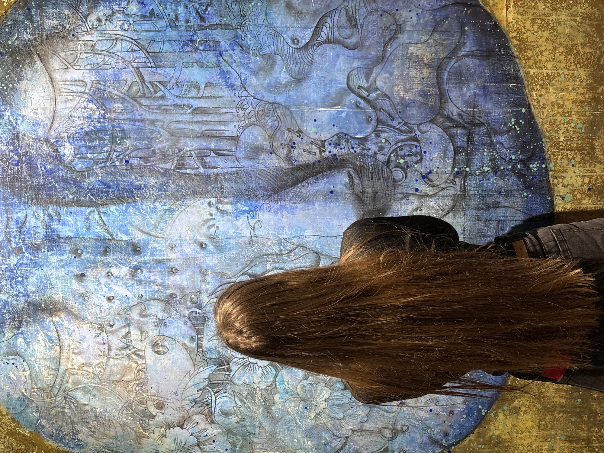 A person with long, brown hair is positioned in front of a large, round artwork. The artwork is a mixed-media piece, predominantly blue and gold, featuring abstract shapes and patterns. The blue areas appear to have a watercolor or wash effect, with various shades of blue and hints of white. The gold is a solid color, suggesting a metallic paint or leafing. The artwork is likely placed on a wall or canvas. The person is wearing a black jacket and jeans.
The exact location in Irkutsk, Russia, cannot be determined from this image alone.