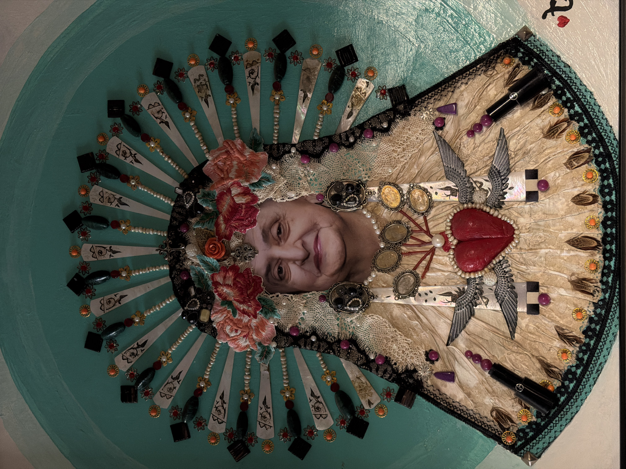 This is an artwork depicting a woman’s face surrounded by various decorative elements. The woman is an older adult with fair skin and a slight smile. Her image is placed at the center of what appears to be an ornate headpiece.
The headpiece is composed of a variety of materials, including lace, pearls, decorative flowers (roses), metallic ornaments, and painted fan-like structures. A large, red, heart-shaped object is centered below the woman’s face, adorned with pearls and flanked by wings. Other objects like lipstick tubes are arranged along the outer edge of the headpiece.
The background is a turquoise-colored surface with subtle variations in shade. A portion of the artwork is framed.
There is no information to indicate that this image relates to the city of Yerevan, Armenia.