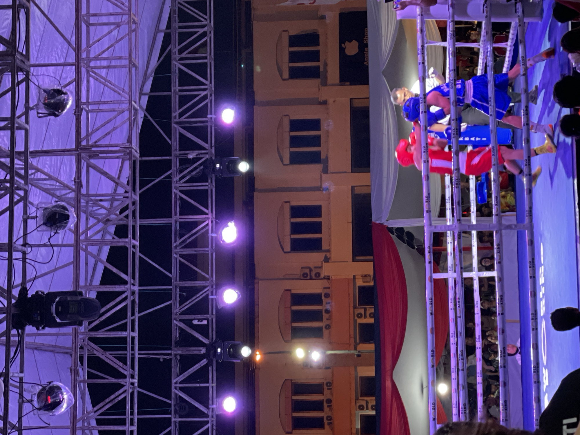 The image depicts a boxing match taking place outdoors at night in Manado, Indonesia. A boxing ring is central to the scene, with two male boxers in action. One boxer, wearing red shorts and a red headguard, is in the process of throwing a punch. The other boxer, in blue shorts and a blue headguard, appears to be blocking. A referee stands behind them, observing the match. Spectators are visible in the background, gathered behind the boxing ring. Above the ring, a metal truss structure supports various lights, some of which are illuminated with purple light. The backdrop features a building with arched windows and air conditioning units, suggesting an urban setting. An Apple Shop sign is visible on the building's facade. The overall atmosphere suggests a professional or semi-professional sporting event under artificial lighting.