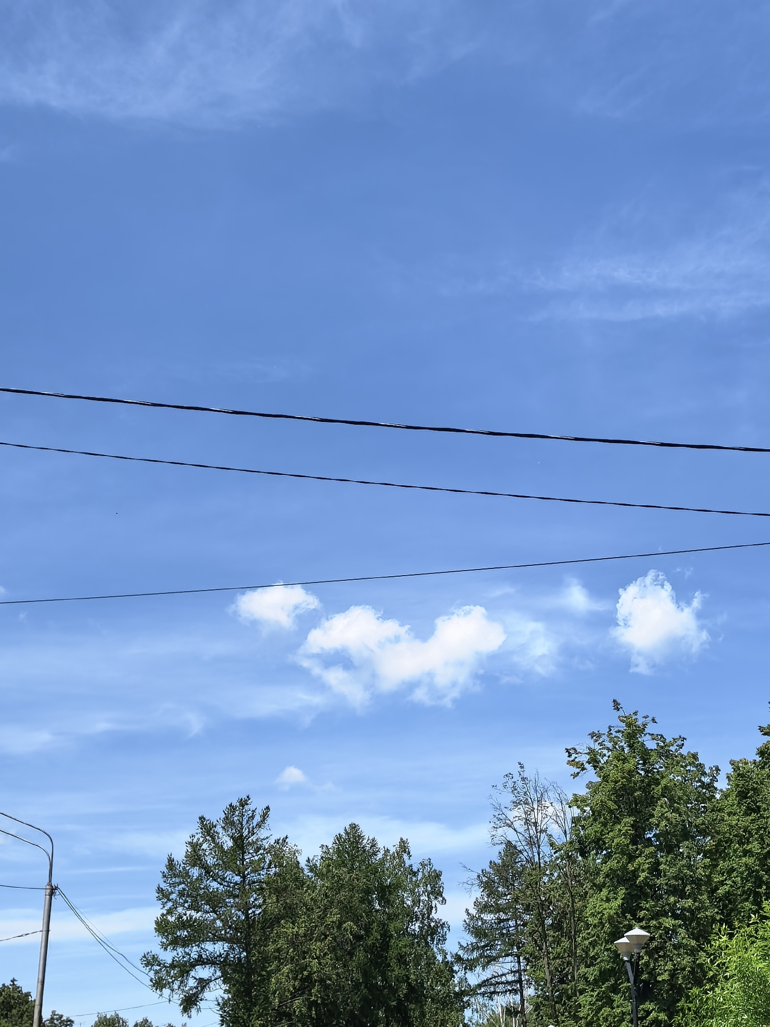 The image captures a daytime scene in Sterlitamak, Russia, featuring a clear blue sky with scattered white clouds. Several horizontal power lines bisect the upper portion of the frame. Below, a row of green trees partially obscures the foreground. A street lamp pole with a dual-light fixture is visible on the left side of the frame. The sky exhibits faint cirrus cloud formations in addition to the cumulus clouds.