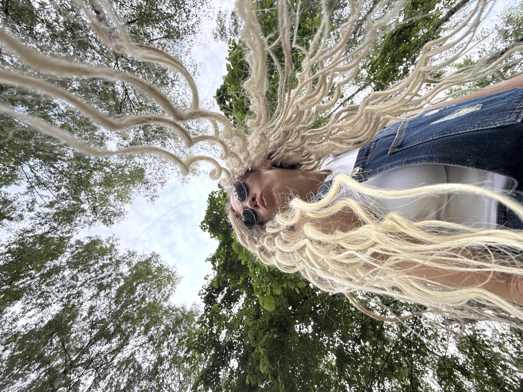 An extreme low-angle, full-body shot captures a young woman with long, blonde dreadlocks looking upwards and slightly to the left, her lips pursed. She is wearing sunglasses, a white tank top, and denim overalls. Her dreadlocks are spread out and appear to be in motion, possibly from wind. The background consists of the green canopy of trees and a cloudy sky, suggesting an outdoor location in a natural setting. The perspective is such that the trees appear to dominate the frame, framing the woman. The image lacks any discernable urban elements of Rusanovo, Russia.