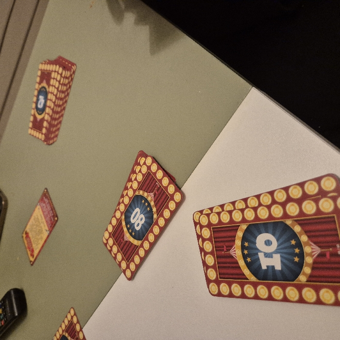 The image shows a collection of cards arranged on a surface with a color transition. One card reads "08" and is decorated with a red background and golden lights around the edges. Another card displays "10" in a similar design with red and gold accents. A stack of cards is also visible along with an additional card with a partially obscured number. A small black object is near the edge of the frame, alongside a cream-colored surface.
