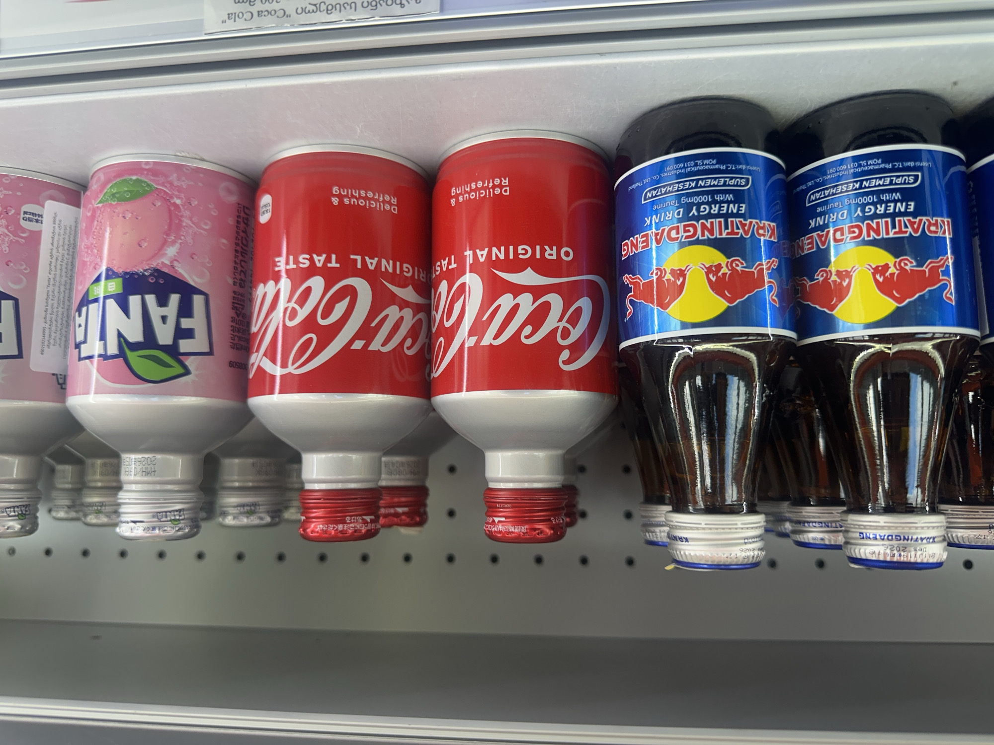 The photograph shows a close-up view of canned and bottled beverages on a shelf, likely in a retail setting, possibly a convenience store or supermarket. The brands visible include Fanta (peach flavored), Coca-Cola, and Krathing Daeng energy drink. The beverages are arranged in a row, with the bottoms of the cans and bottles facing the viewer. The bottoms display details such as expiration dates and manufacturer information, with some text in Asian languages and also in Indonesian. The shelf is a plain grey, utilitarian fixture. The photograph provides no explicit information to suggest that this location is Zeda Bashi, Georgia.