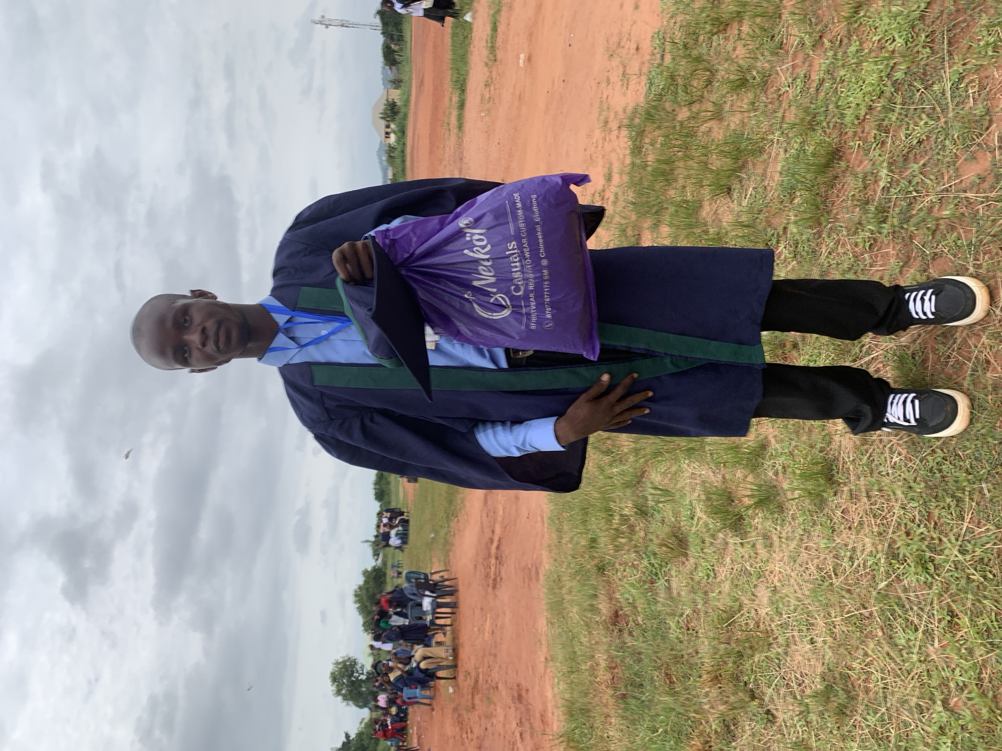 A man stands outdoors in a grassy field under a cloudy sky. He wears a graduation gown over a light blue shirt and black pants, paired with black and white sneakers. He holds a purple shopping bag with "Nerkol Casuals" printed on it. The bag also displays contact information: "STRIETWEAR, READY-TO-WEAR.CUSTOM-MADE" and "07077677175 SM: @ Chineekel Clothing." In the background, a gathering of people can be seen, possibly seated in chairs. Red dirt paths are visible between the grassy areas. This image could have been captured in Nsugbe, Nigeria, based on the information in the bag.